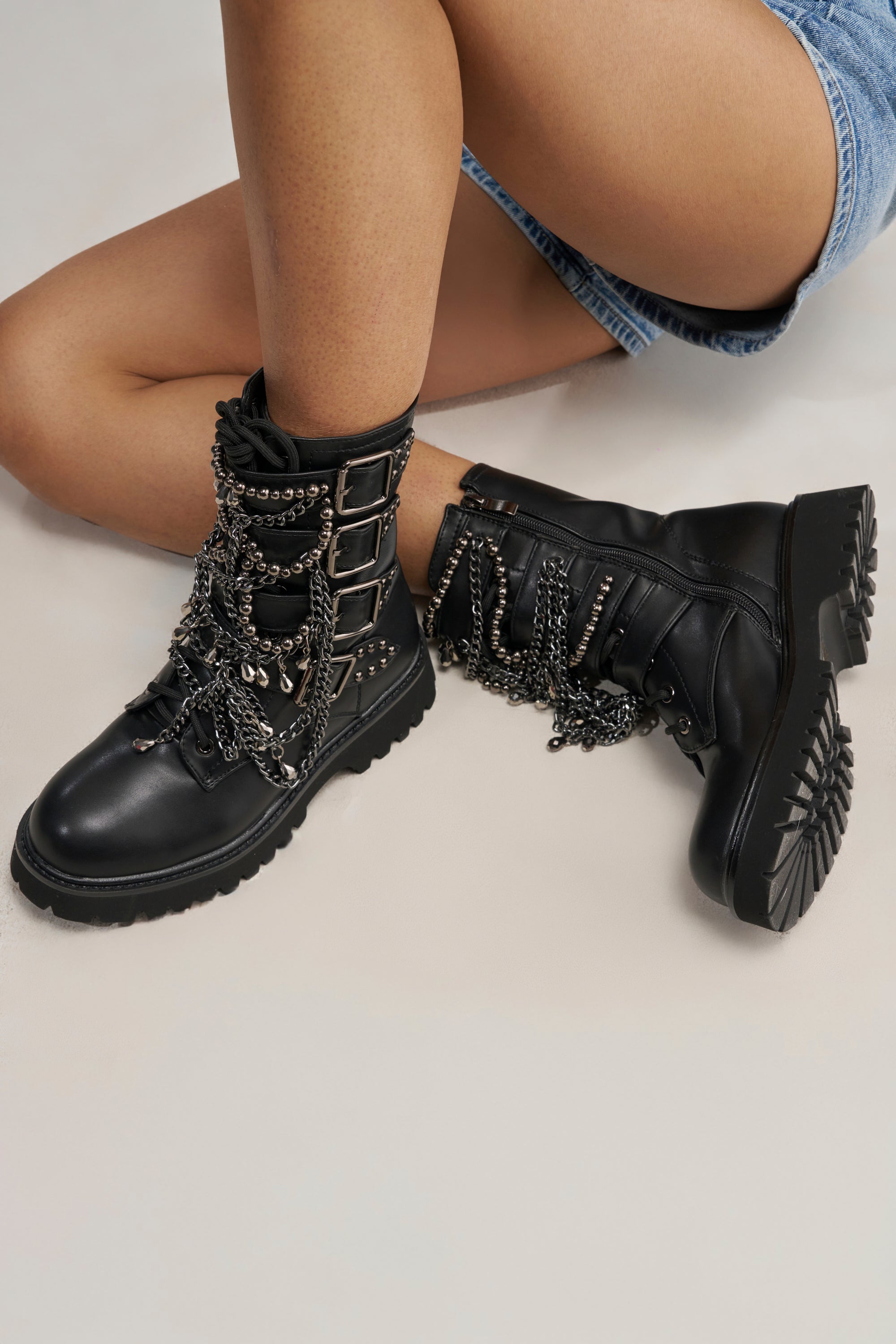 Wearing the AZALEA WANG ERIN EMBELLISHED COMBAT BOOTIE—black faux leather boots adorned with chains and buckles—a person sits with bent legs on a light surface, styled with denim shorts.