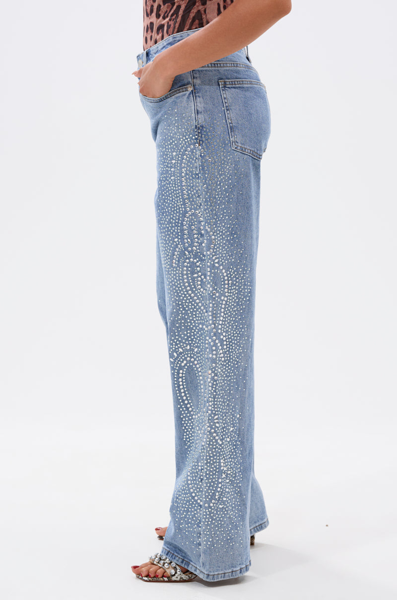 A woman models the DREAMING OF YOU WIDE LEG DENIM, featuring light blue high-rise jeans with rhinestone side embellishments. She stands sideways, hand in pocket, styled with open-toed heels and an animal print top for a chic vibe. 3