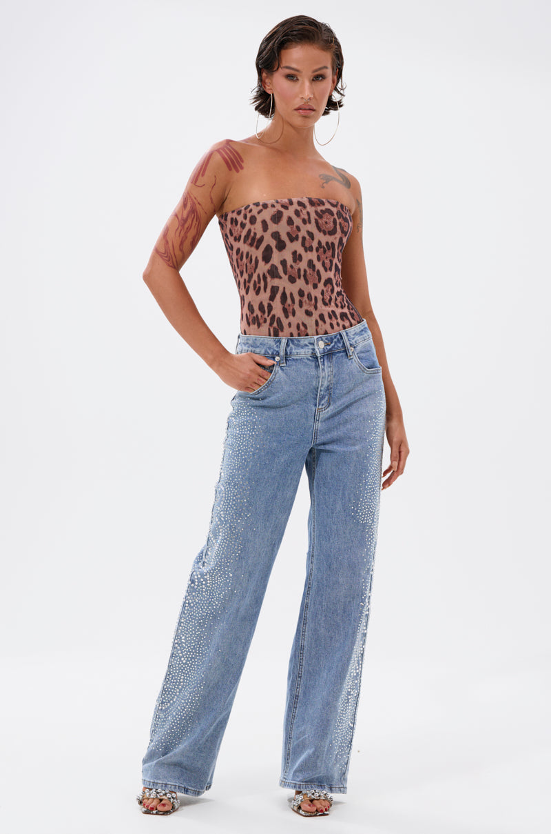 A person poses confidently in DREAMING OF YOU WIDE LEG DENIM, featuring high-rise, wide legs and rhinestone side embellishments, paired with a strapless leopard-print top. Their look is completed with wet-look short hair and visible arm tattoos for an edgy vibe. 6