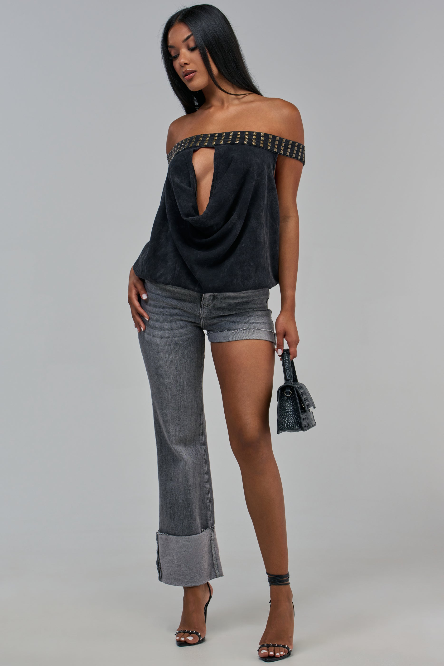 A person wears a black off-the-shoulder top with a studded neckline and a cutout, paired with BOTH SIDES OF ME DENIM IN BLACK, which feature an asymmetrical leg design, while holding a small handbag.