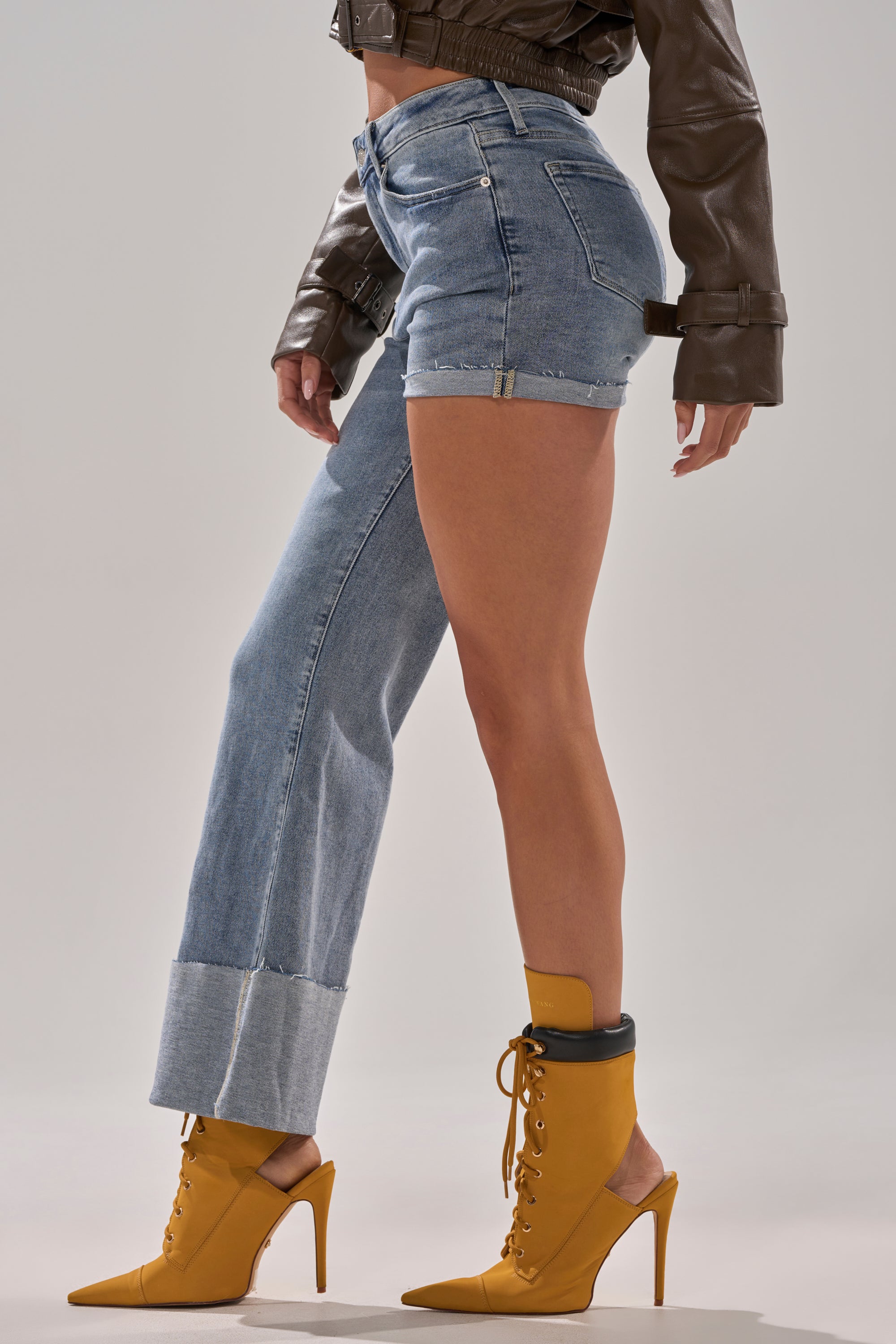 Wearing the BOTH SIDES OF ME DENIM—with one leg full-length and the other cropped—paired with a brown leather jacket and mustard yellow lace-up boots, a confident figure poses against a plain background.