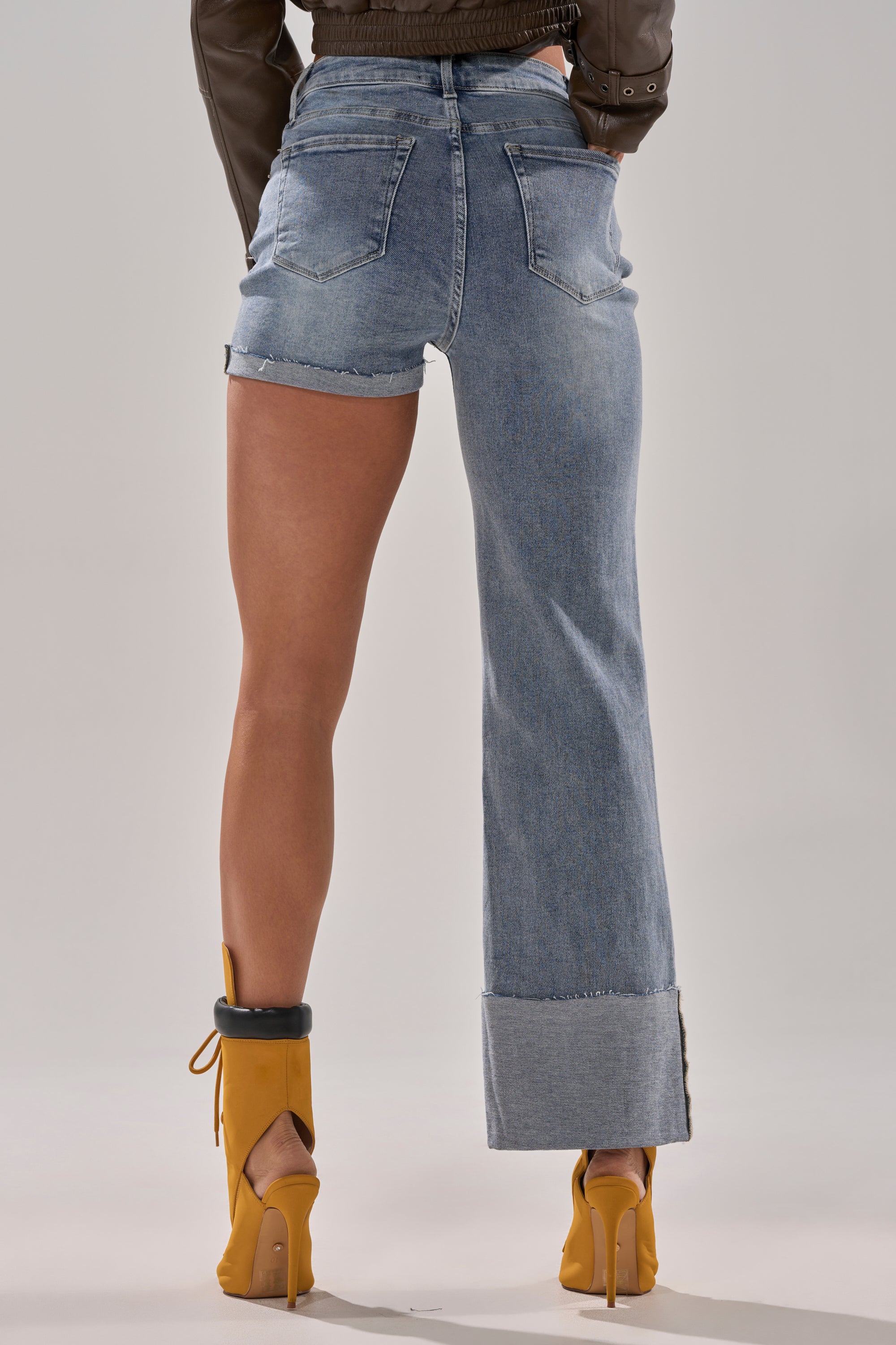Someone wears the BOTH SIDES OF ME DENIM—a pair of light-wash high-waisted jeans with mismatched hem (one side shorts, one full-length)—styled with yellow heels and a brown jacket against a plain, neutral background.