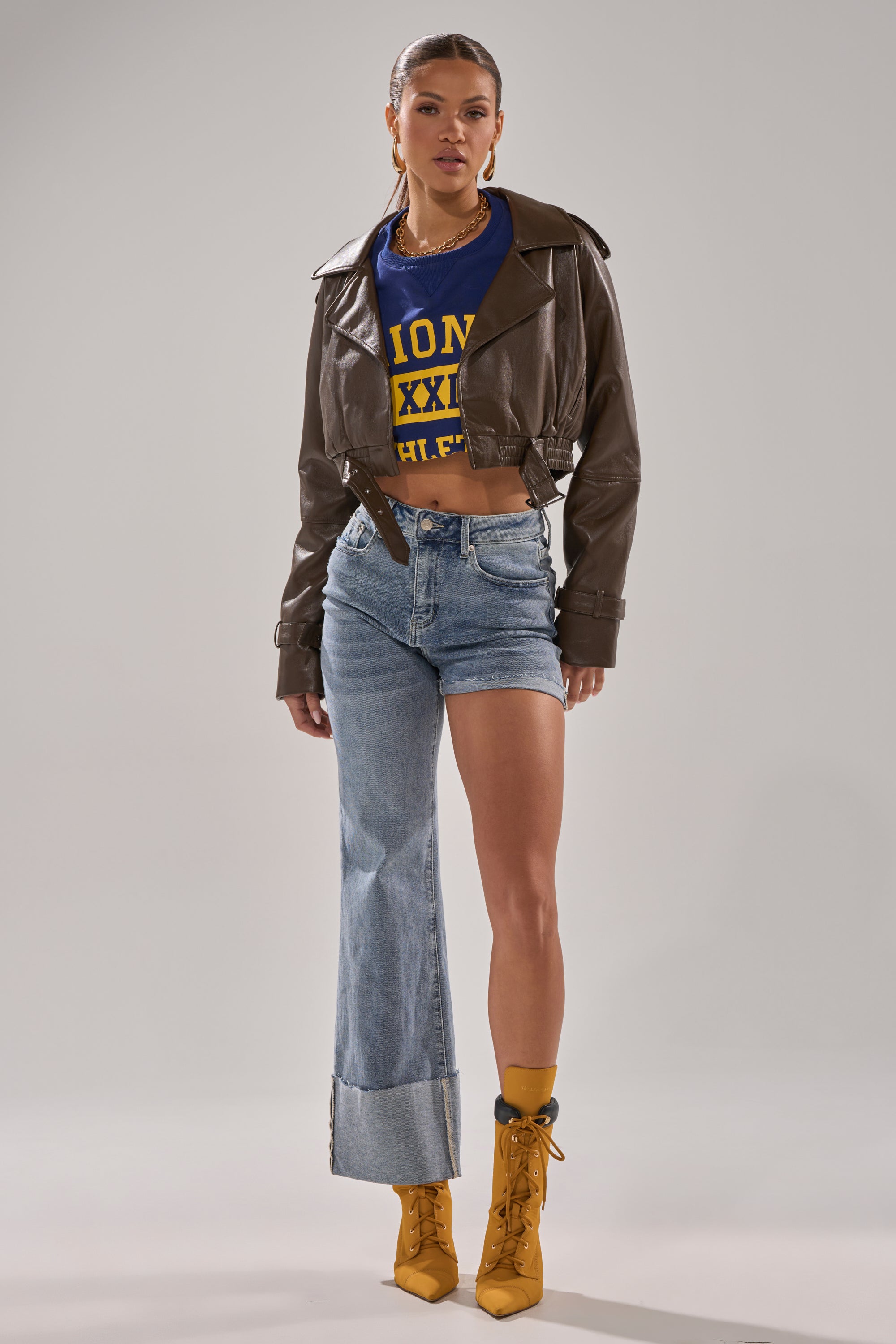 A woman poses against a plain background wearing BOTH SIDES OF ME DENIM jeans featuring one shorts leg and one full-length leg, styled with a cropped brown leather jacket, a blue and yellow graphic crop top, and yellow lace-up heeled boots.