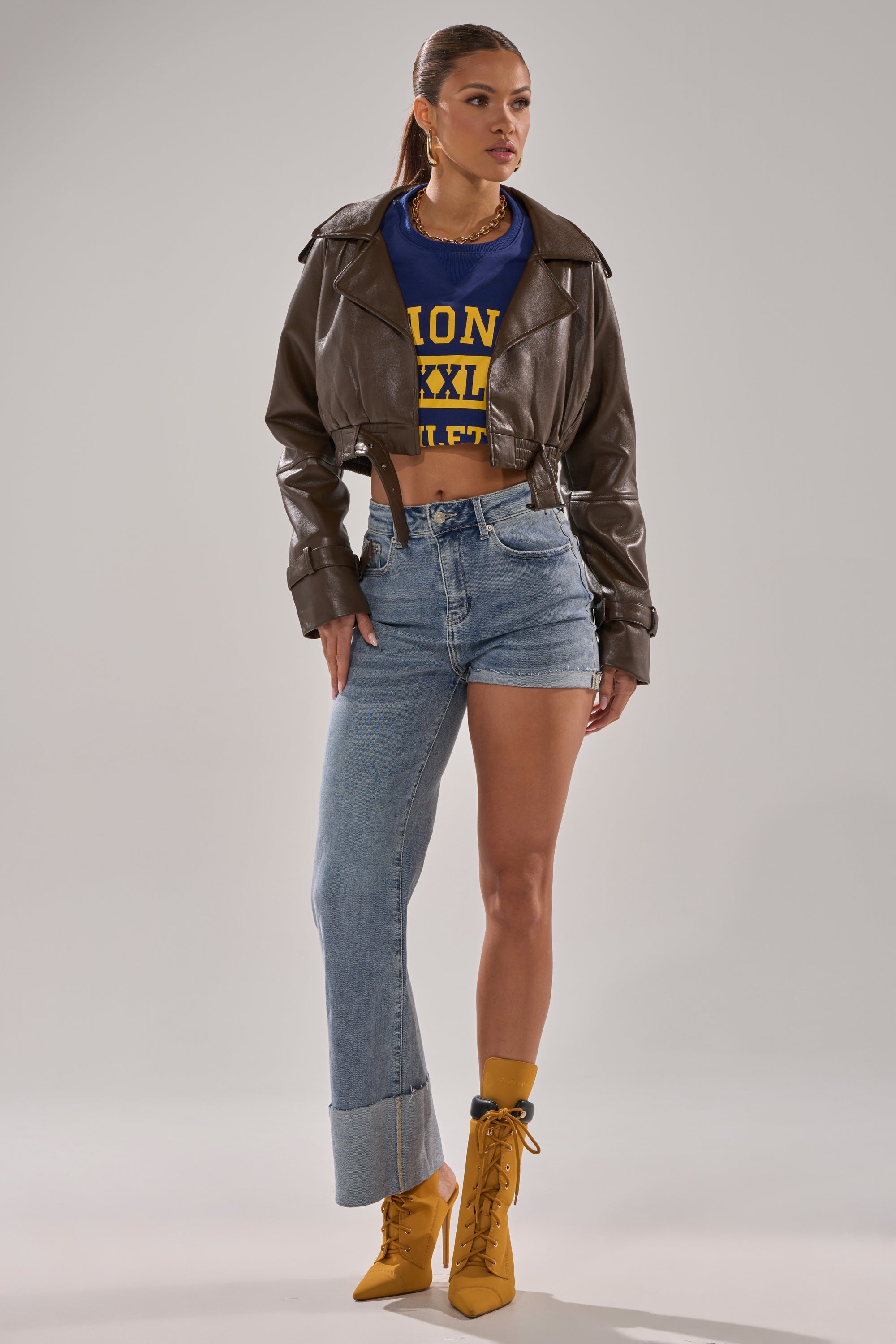 A woman wears the BOTH SIDES OF ME DENIM—high-rise jeans with one full-length cuffed leg and one cut-off short leg—styled with a brown cropped leather jacket, blue and yellow crop top, and yellow lace-up heels over matching socks.