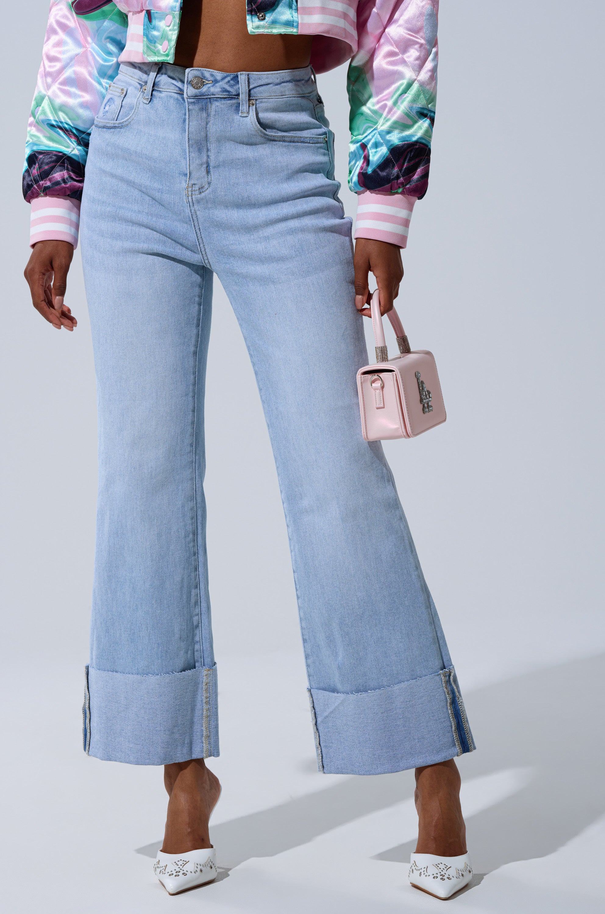 A person wearing LOVE ME MORE RELAXED DENIM jeans (light blue, high-rise, wide-leg with large cuffs), a shiny multicolored cropped jacket, white pointed heels, and carrying a small pink handbag stands against a plain light gray background.