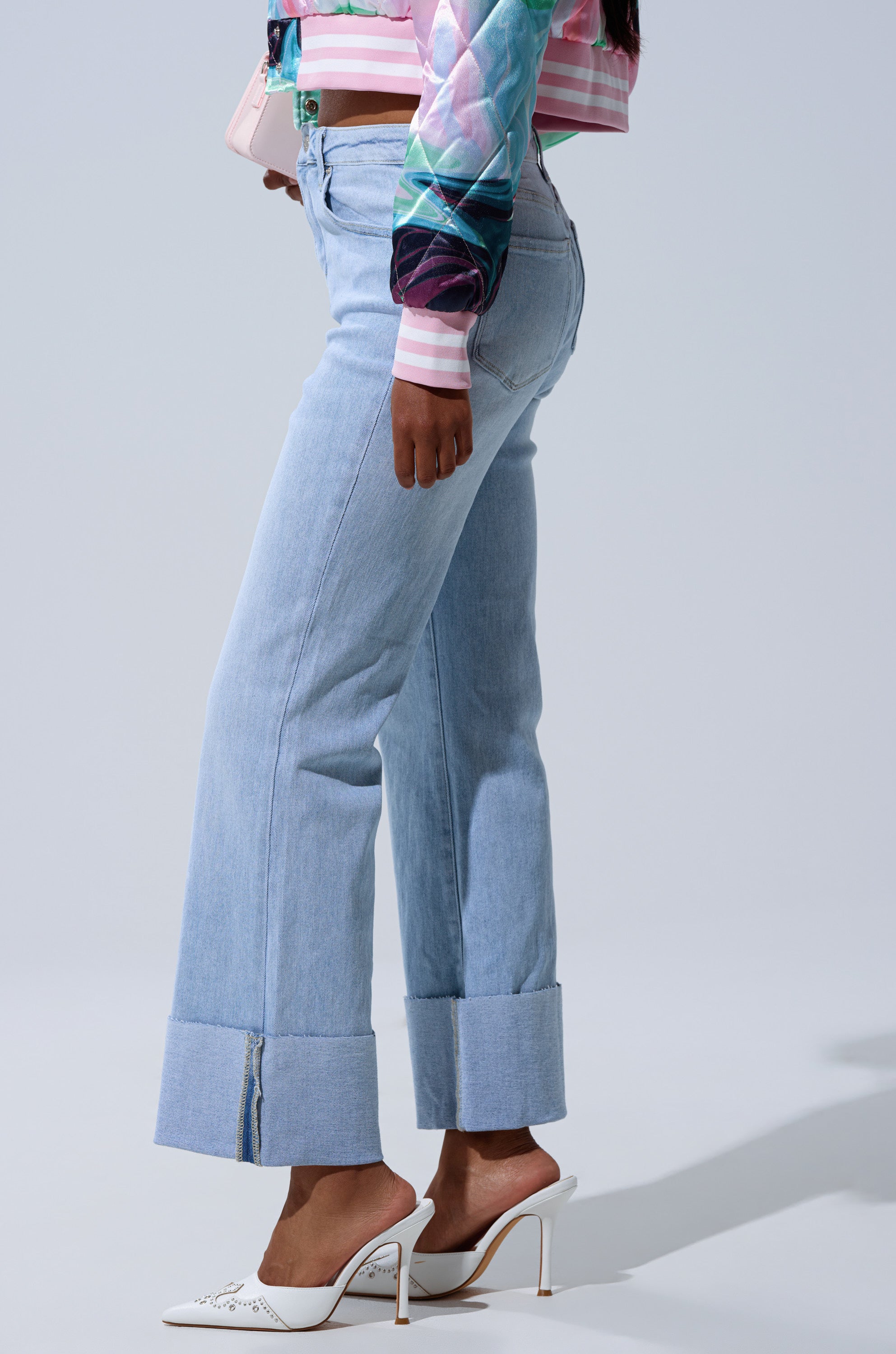 A woman in profile wears the LOVE ME MORE RELAXED DENIM, cuffed at the ankle, paired with a colorful patterned jacket and white open-back heeled shoes against a plain light background.