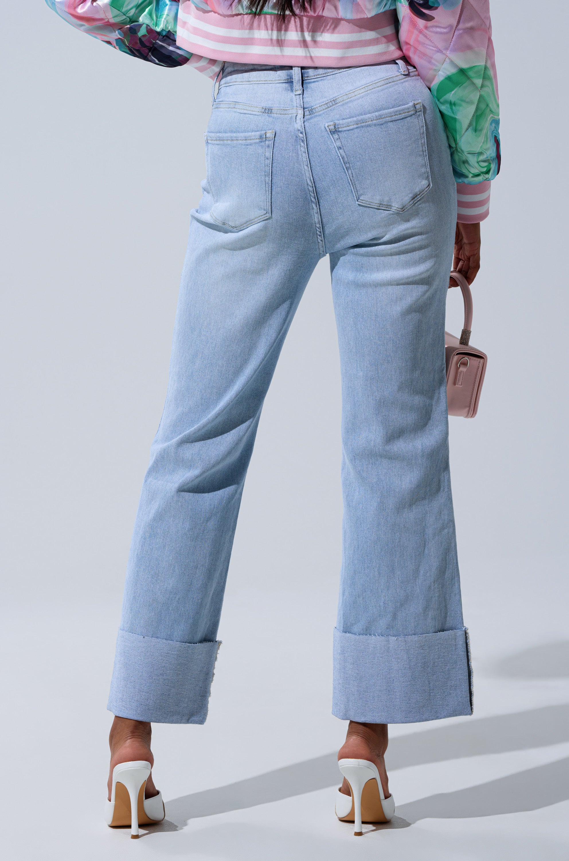 A person stands facing away against a plain light background, wearing LOVE ME MORE RELAXED DENIM cuffed high-rise straight-leg jeans, white heels, a colorful top, and holding a small pink handbag by their side.