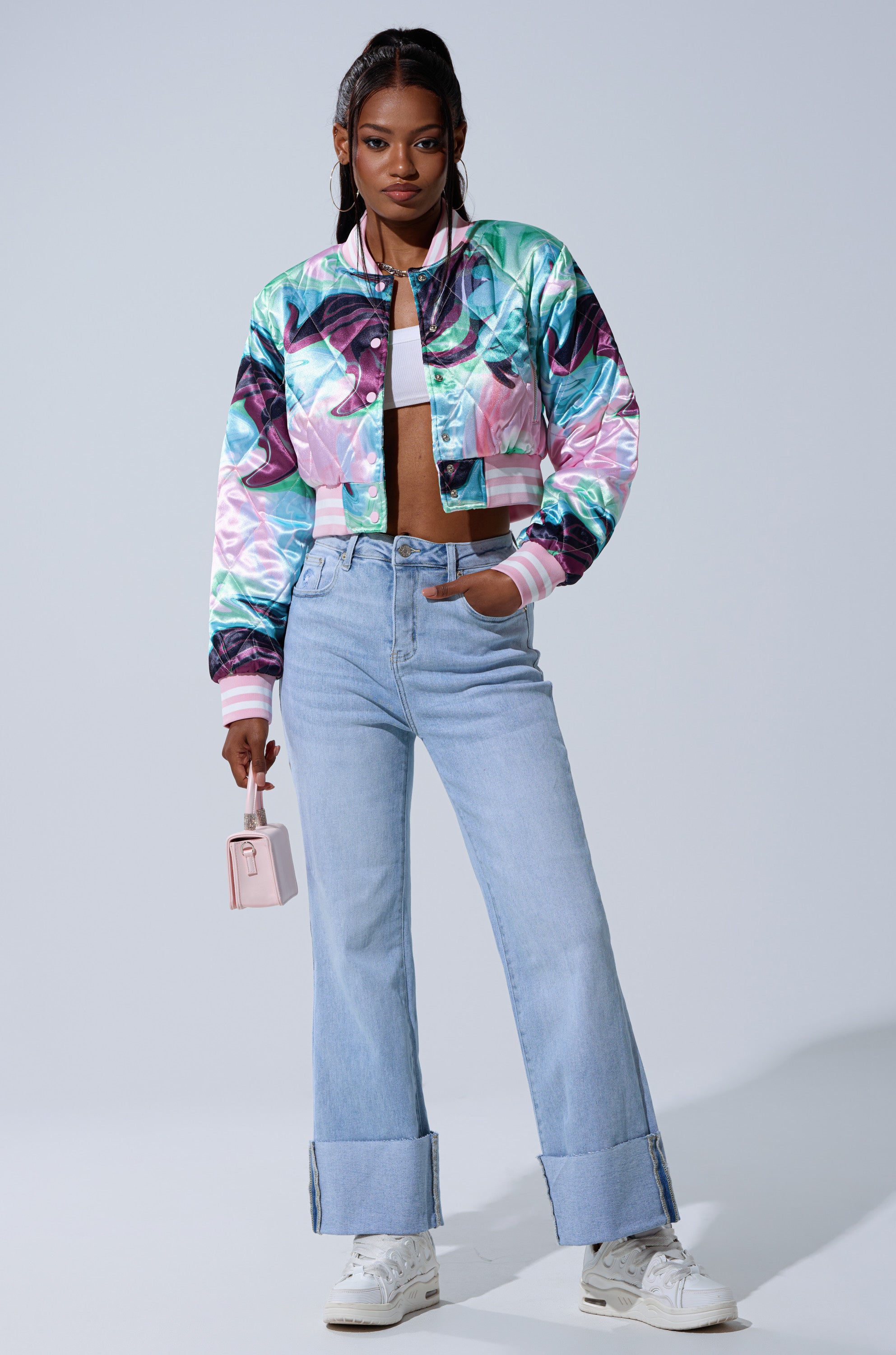 A woman poses confidently in LOVE ME MORE RELAXED DENIM jeans with large cuffs, a colorful shiny cropped jacket, white crop top and sneakers, holding a small pink handbag against a plain background.
