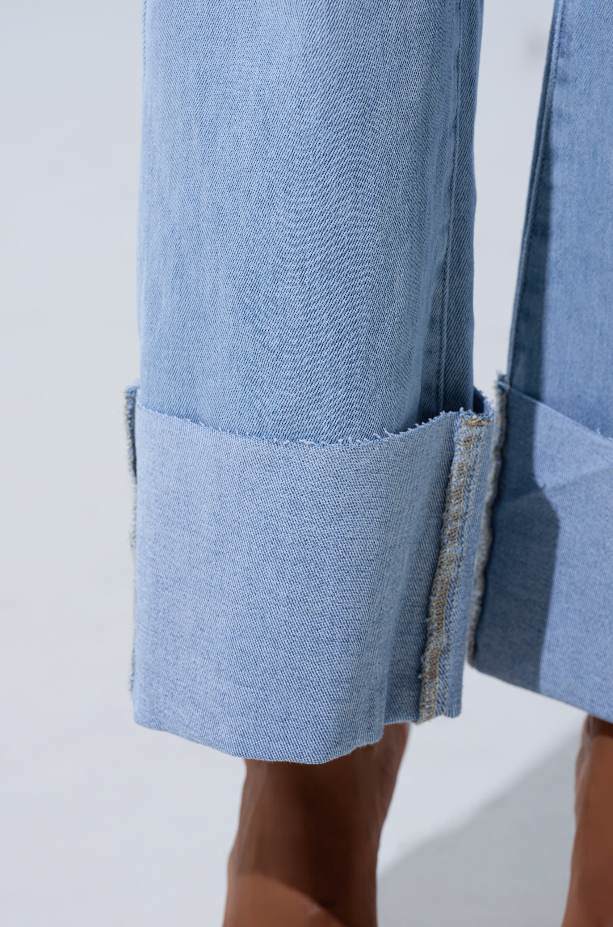 Close-up of the LOVE ME MORE RELAXED DENIM: light-wash, high-rise straight-leg jeans featuring wide, double-folded raw-edged cuffs at the ankles. Set against a plain background with part of a bare foot visible below the cuff.