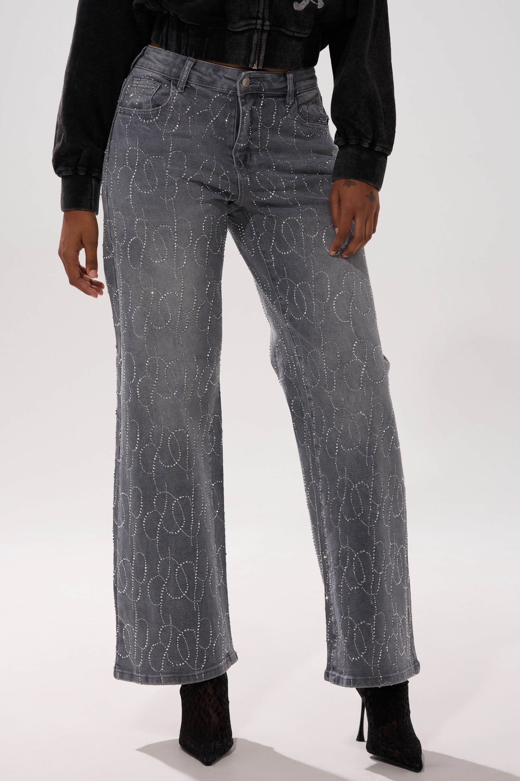 A person wears the PERFECT LIKE ME EMBELLISHED DENIM—high-waisted, wide-leg gray jeans with a subtle circular pattern—styled with a black cropped jacket and black heeled boots.