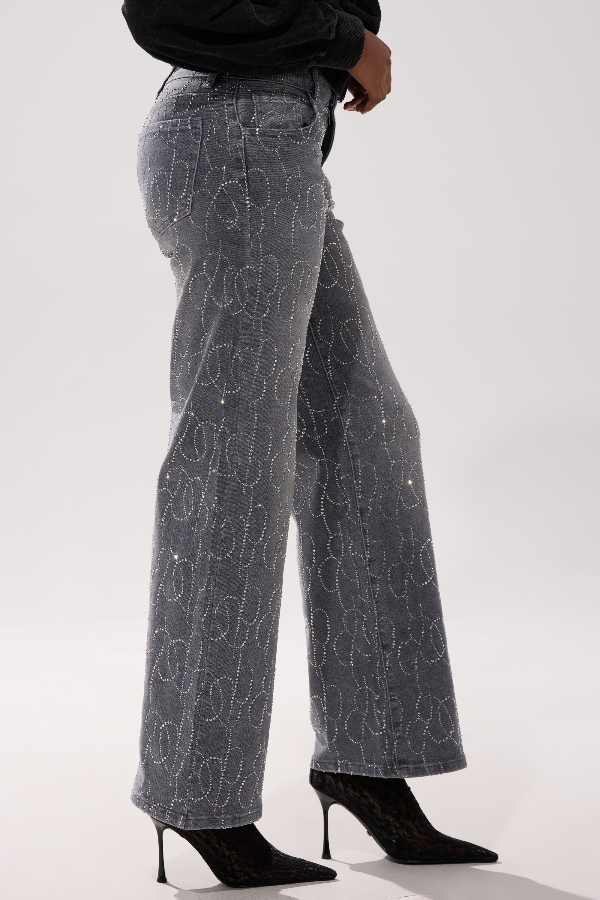 A person wearing the PERFECT LIKE ME EMBELLISHED DENIM—high-rise, straight-leg gray jeans with decorative stitching—pairs them with black pointed-toe heels and a dark top, standing against a plain light background.