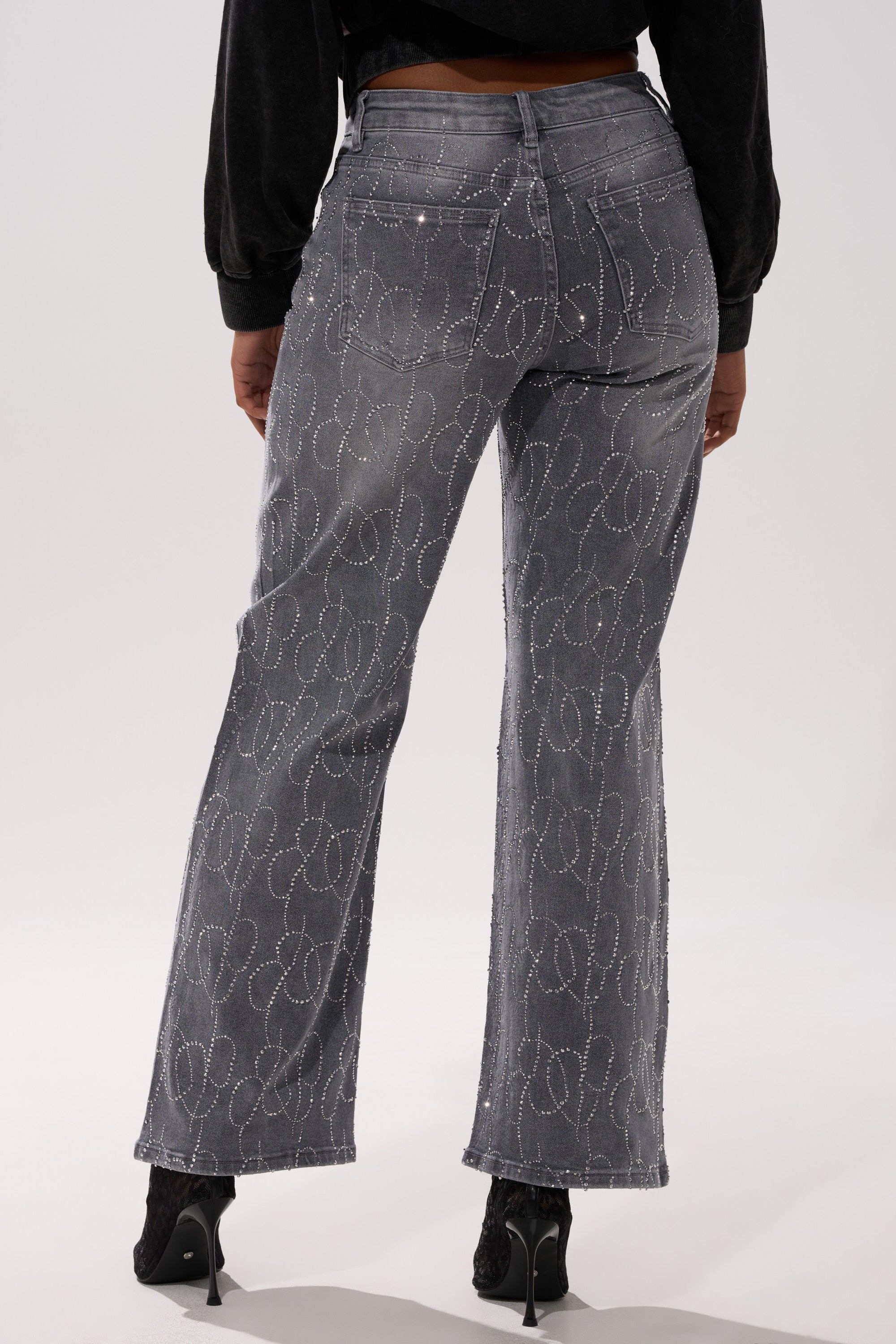 A person stands facing away, wearing the PERFECT LIKE ME EMBELLISHED DENIM jeans with a subtle faded wash and white stitched patterns, paired with black shoes and a black long-sleeve top.