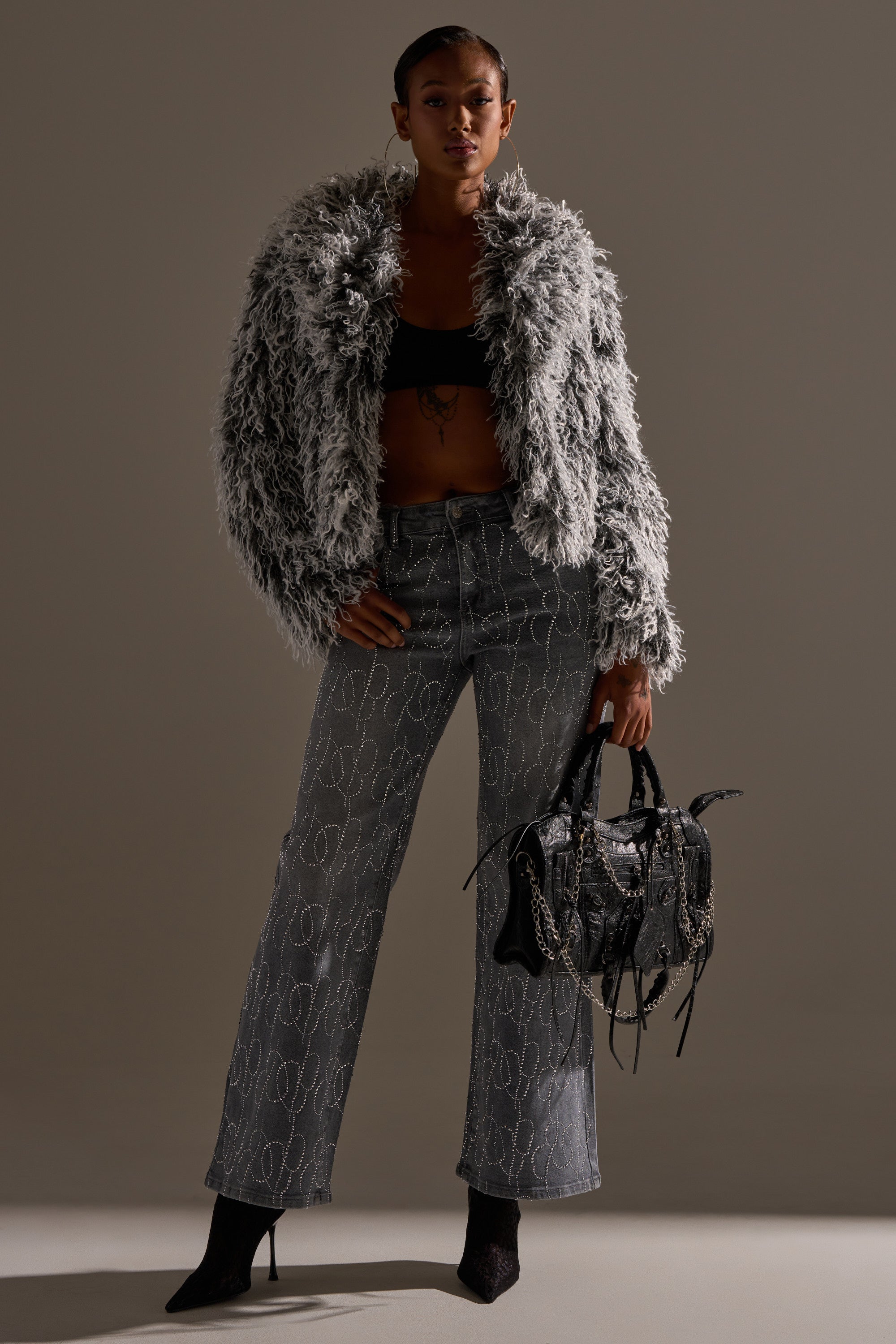 A woman stands confidently in the PERFECT LIKE ME EMBELLISHED DENIM, styled with a gray faux fur jacket, black crop top, black boots, and a fringed chain-detail handbag. She poses against a plain, softly lit background.