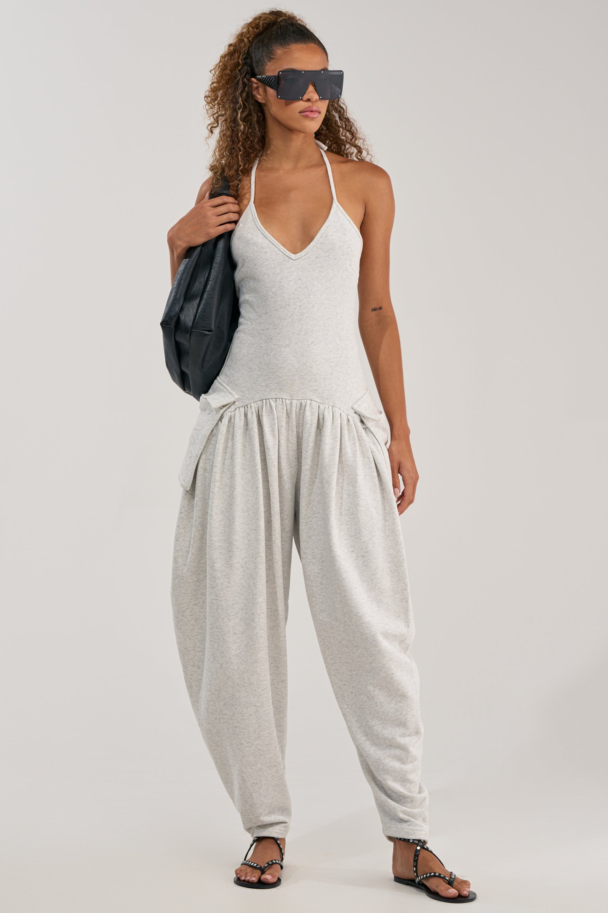 Wearing the COZY AF KNIT JUMPSUIT with a deep neckline and loose pants, a woman in oversized sunglasses and black sandals stands against a plain background, carrying a black bag on her shoulder.
