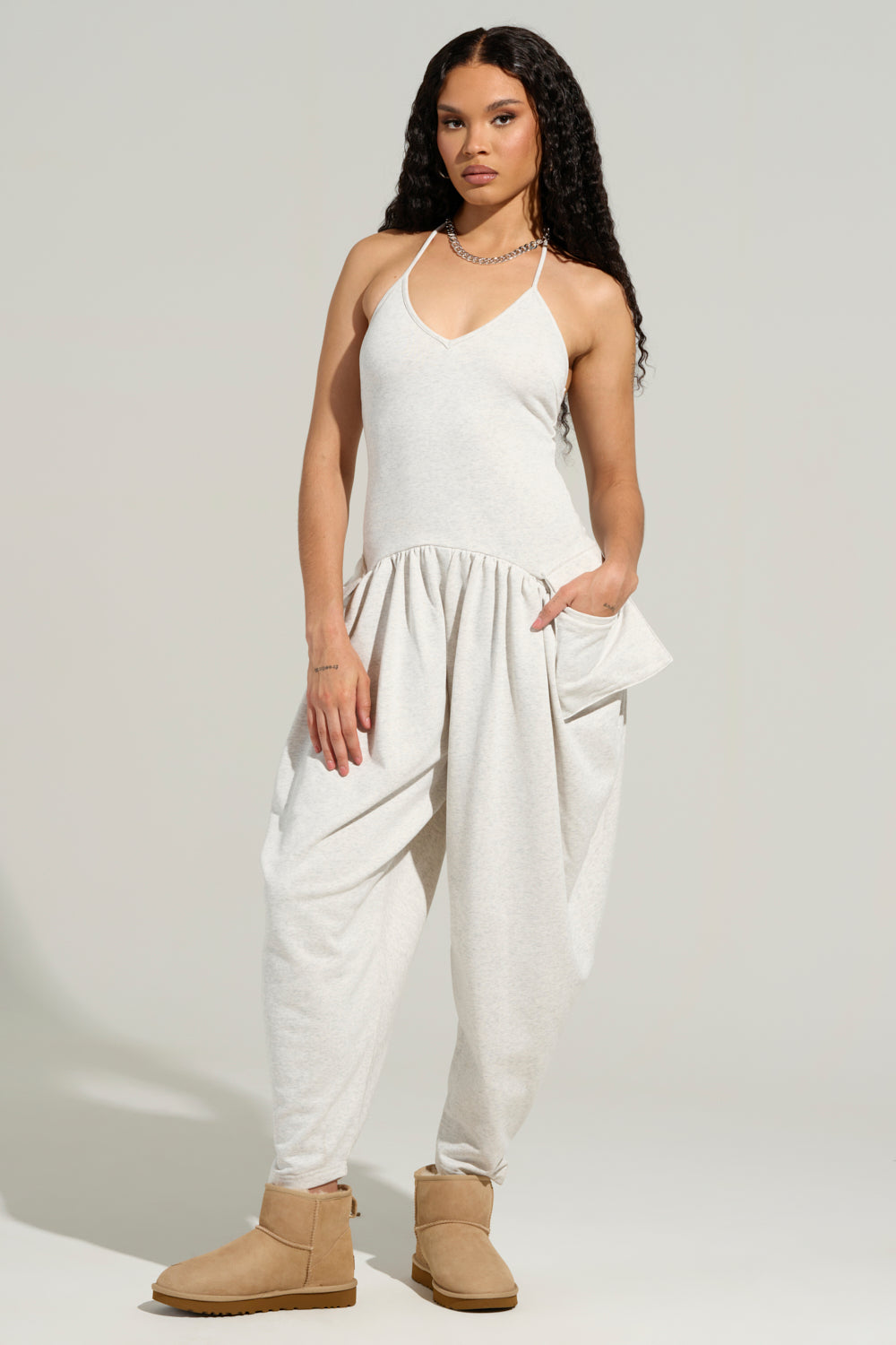 A person with long curly hair is wearing the COZY AF KNIT JUMPSUIT in light gray, which features a sleeveless, backless silhouette and wide legs. They are standing against a plain background, complemented by tan ankle boots and a necklace.