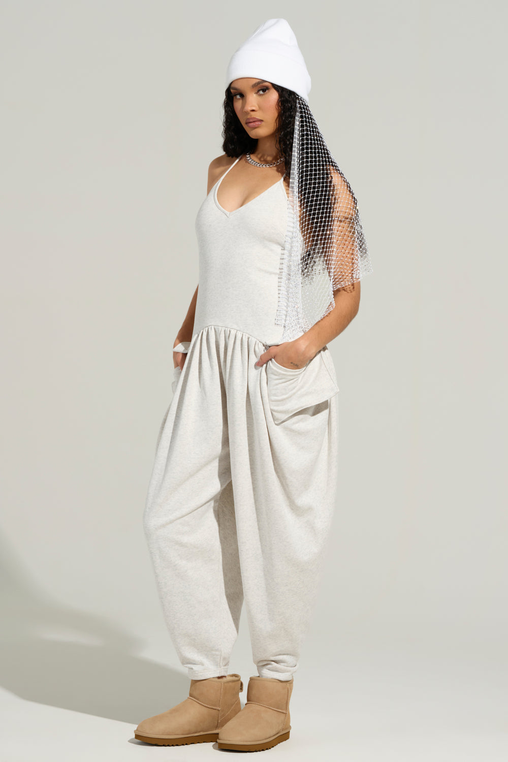 A person exudes confidence against a neutral background, flaunting a white beanie, a long mesh veil, and the COZY AF KNIT JUMPSUIT in beige, featuring pockets and wide pants. Their tan boots perfectly complete the look.