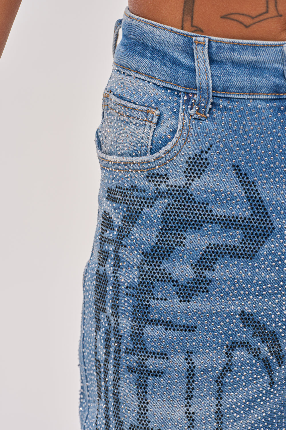 Close-up of the ITS MAGIC RHINESTONE STRAIGHT LEG DENIM: blue denim jeans with black graphic designs and sparkling rhinestones. A visible tattoo above the waistband heightens their edgy, fashion-forward appeal. 9