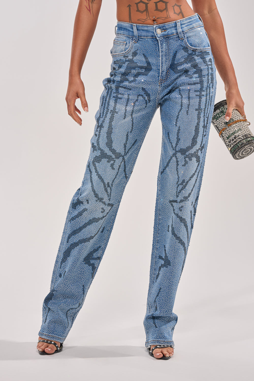 A person wears ITS MAGIC RHINESTONE STRAIGHT LEG DENIM—high-waisted, blue, marble-effect jeans—in street style, paired with open-toe heels and holding a cylindrical beaded clutch. Only their torso and legs are shown. 2