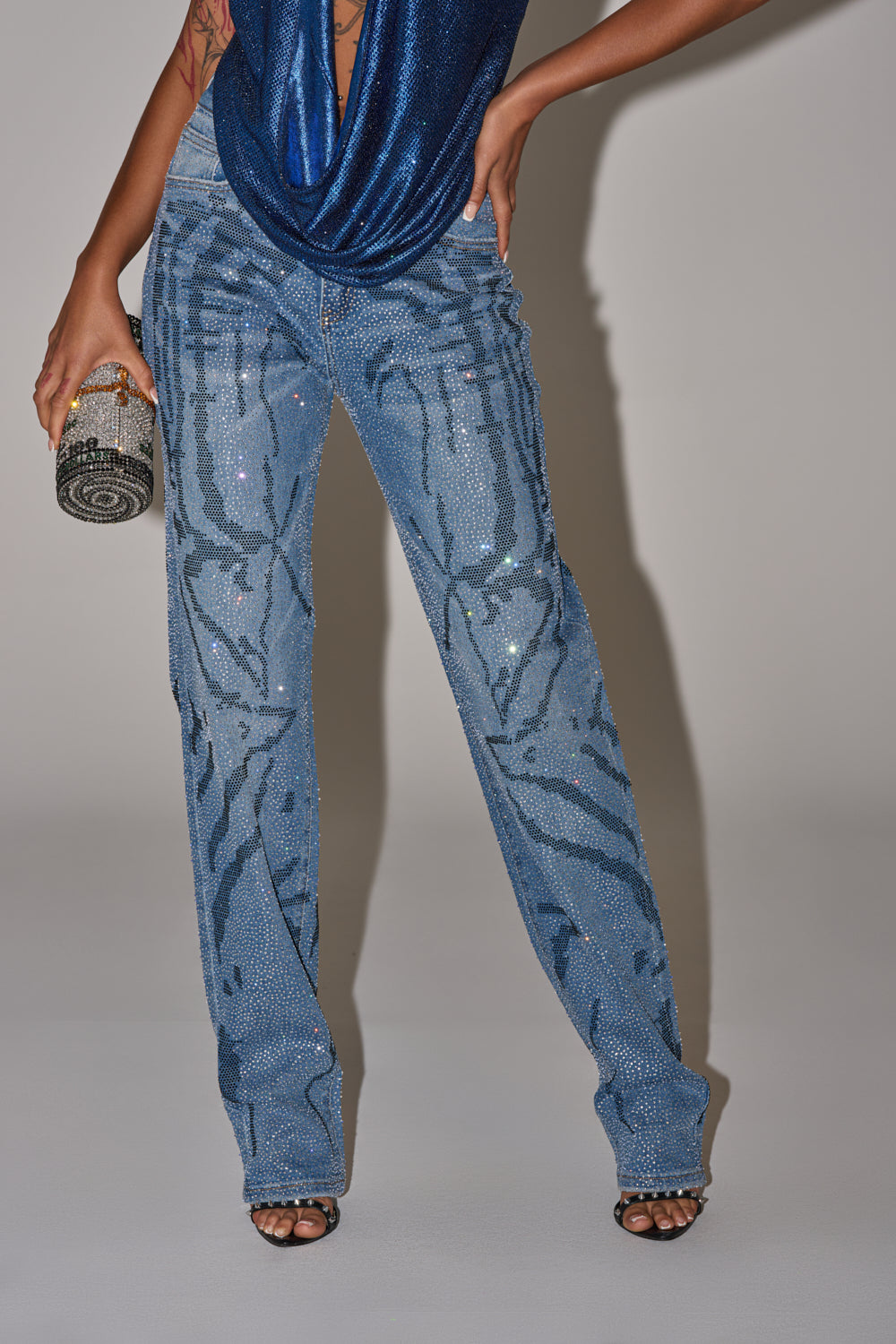 A person wearing ITS MAGIC RHINESTONE STRAIGHT LEG DENIM featuring a wavy rhinestone pattern, paired with a shiny blue top, black strappy heels, and holding a small rhinestone-studded clutch. Image is cropped at the shoulders. 0