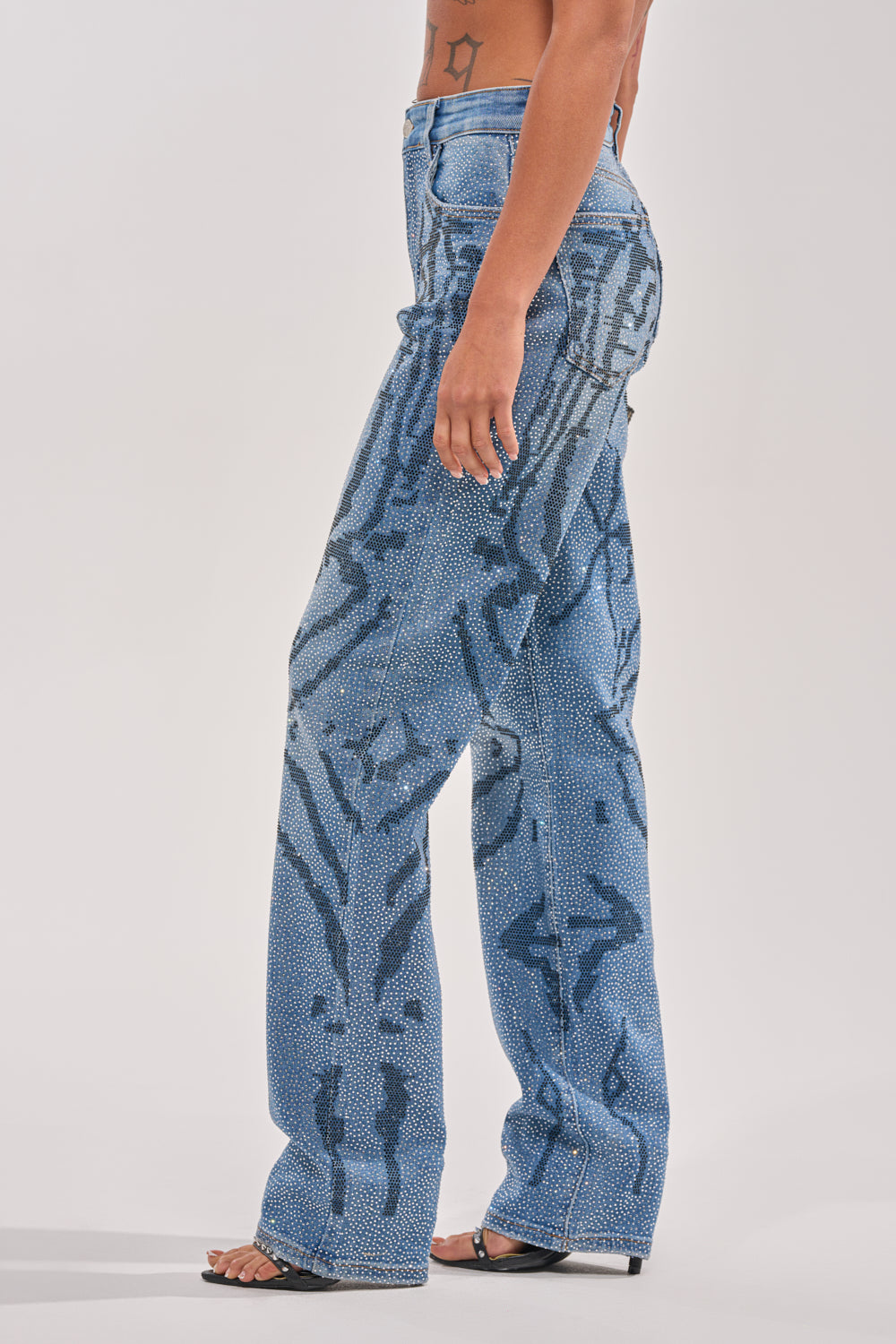 A person stands in profile wearing the ITS MAGIC RHINESTONE STRAIGHT LEG DENIM, designed with a bold, abstract black pattern and high-waisted, loose fit; paired with black open-toe heels, their midriff and tattoo are partly visible. 4