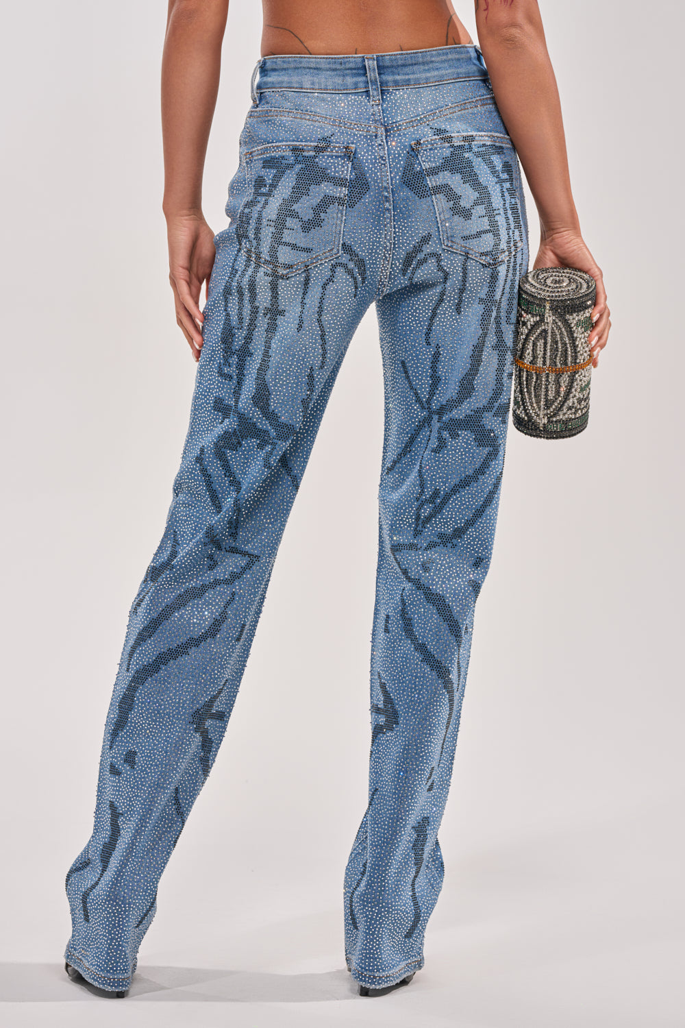 A person faces away, wearing ITS MAGIC RHINESTONE STRAIGHT LEG DENIM with a bold zebra pattern and holding a sparkling, beaded cylindrical clutch against a plain, light background. 5