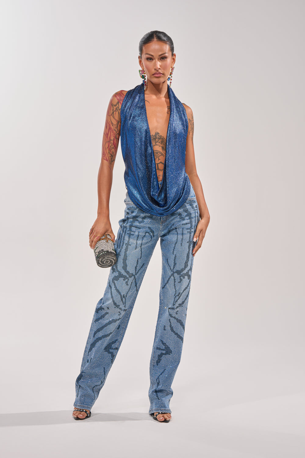A woman stands confidently in ITS MAGIC RHINESTONE STRAIGHT LEG DENIM, a deep blue sleeveless draped top, and high heels, accessorized with statement earrings, a decorative clutch, and visible tattoos on her arms and chest. 7