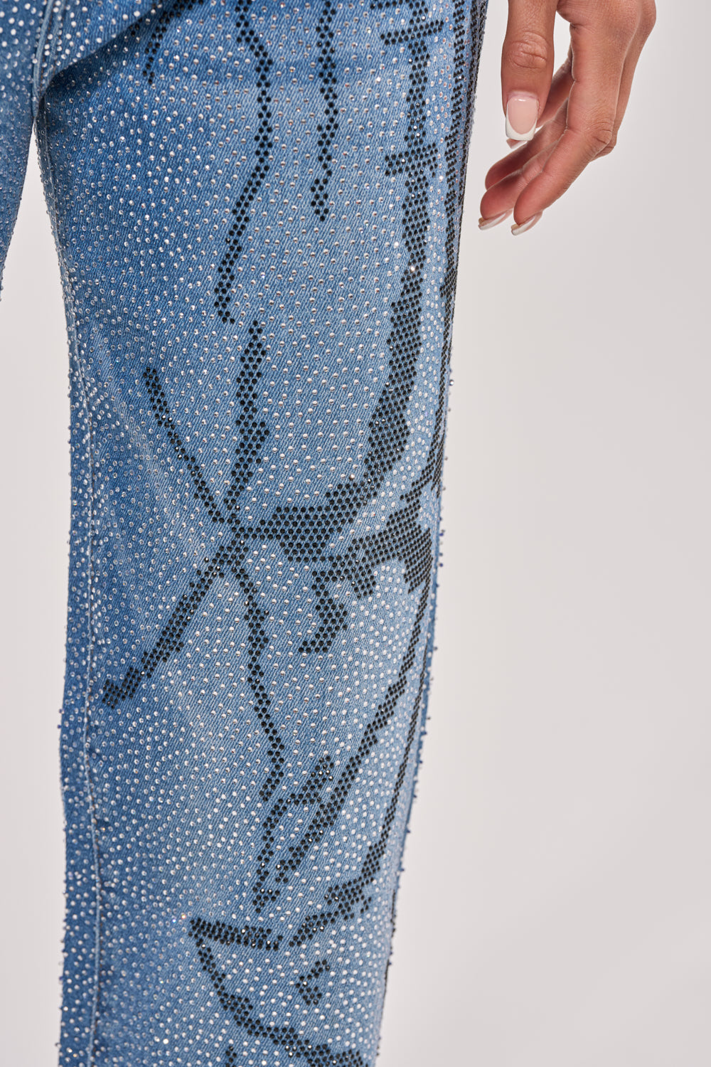 A close-up shows a hand near the hip wearing the ITS MAGIC RHINESTONE STRAIGHT LEG DENIM: blue street-style jeans adorned with sparkling rhinestones and a black abstract line pattern, set against a plain light background. 8