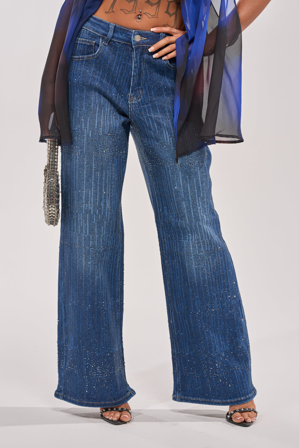 A person wears the ITS ELECTRIC RHINESTONE WIDE LEG DENIM, featuring high-waisted, wide-leg blue jeans with sparkling rhinestones, paired with black open-toe heels and a sheer black and blue top. Only the lower body is visible against a light background. 0