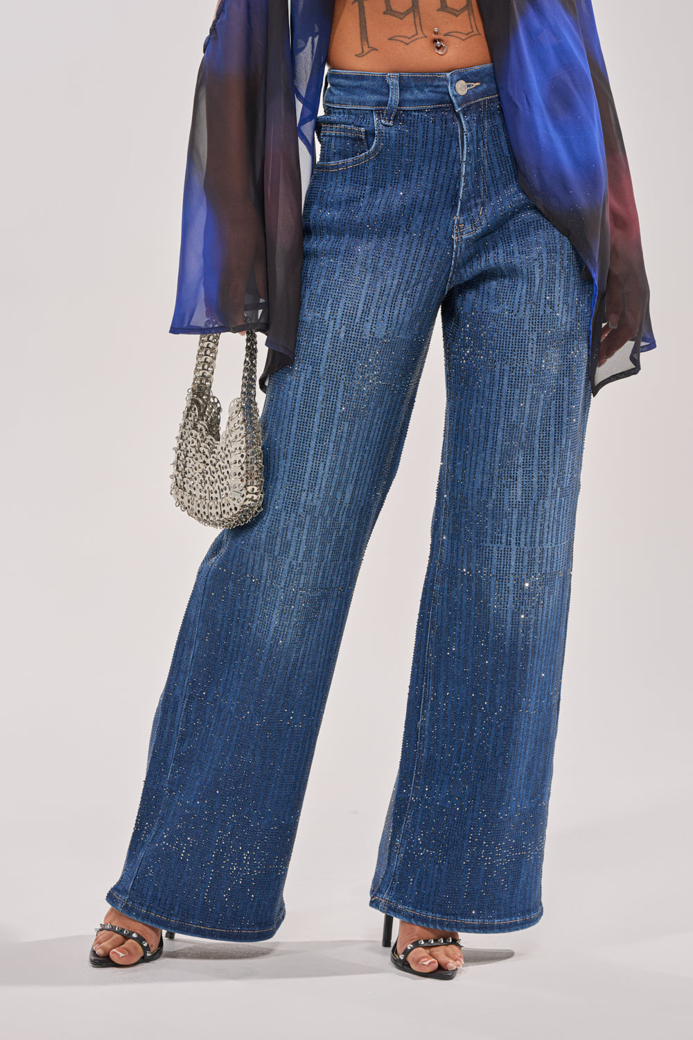 A person in ITS ELECTRIC RHINESTONE WIDE LEG DENIM, paired with a sheer flowy top, heeled sandals, and a small silver beaded handbag. 1