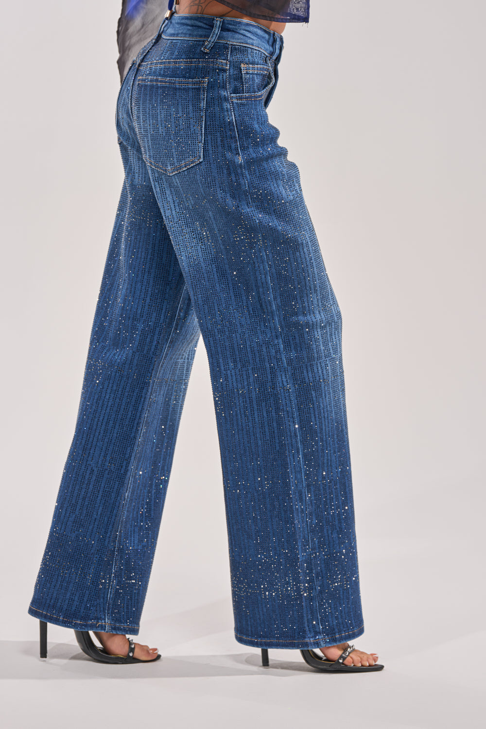Wearing the ITS ELECTRIC RHINESTONE WIDE LEG DENIM, this person stands against a plain light background, showcasing Y2K style with sparkly embellishments and black open-toe heeled sandals. 2