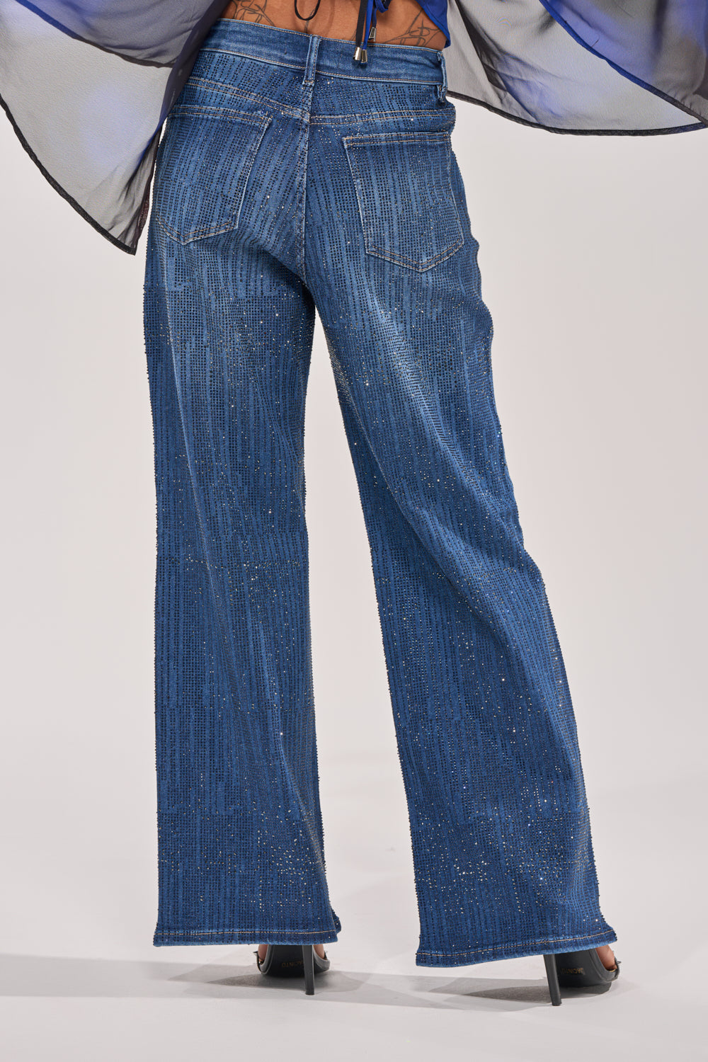 From the back, a person models the ITS ELECTRIC RHINESTONE WIDE LEG DENIM in sparkling blue Y2K style with visible pockets, subtle shimmer, and a sheer, flowing accent at the waist, paired with black high-heeled sandals. 3