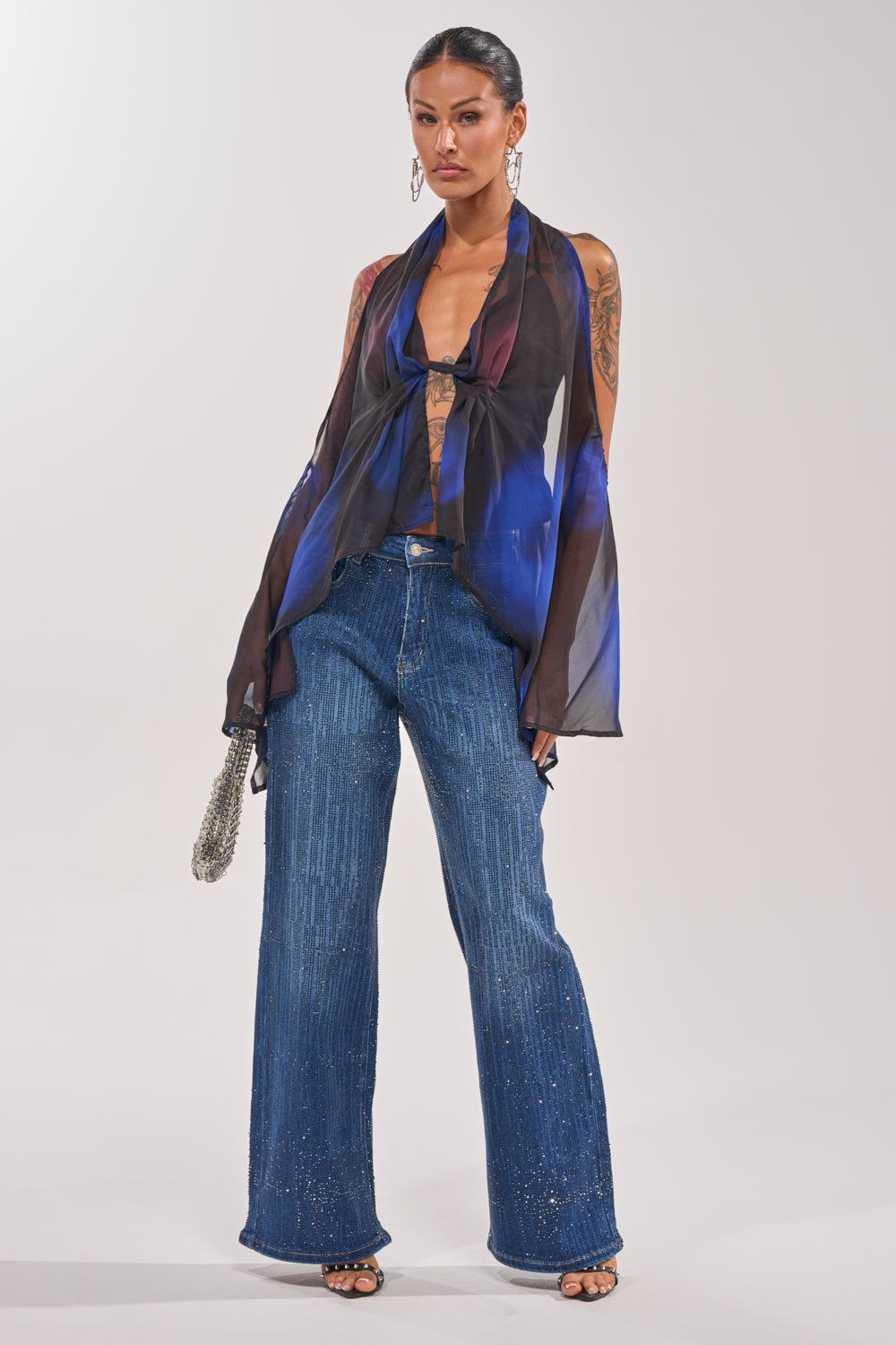 A woman models ITS ELECTRIC RHINESTONE WIDE LEG DENIM, styled Y2K with a dark sheer halter top, hoop earrings, and a small silver bag, posing against a plain white background. 4