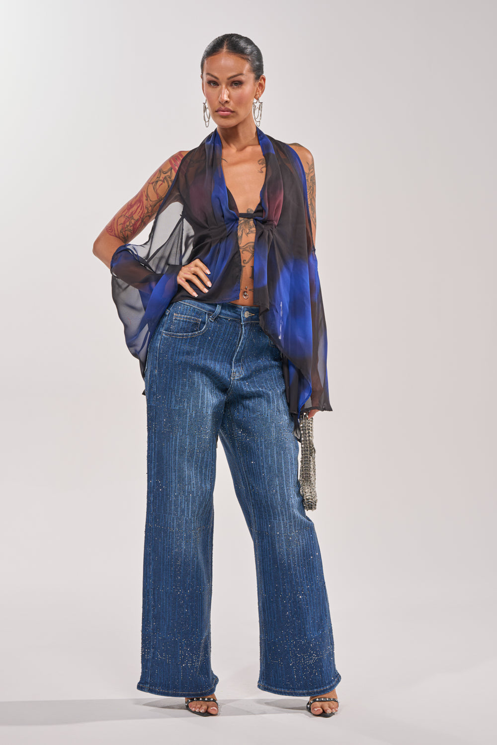A woman exudes Y2K vibes in ITS ELECTRIC RHINESTONE WIDE LEG DENIM, paired with a sheer blue and black waist-tied top over a bralette, open-toe heels, statement earrings, and arm tattoos. 5
