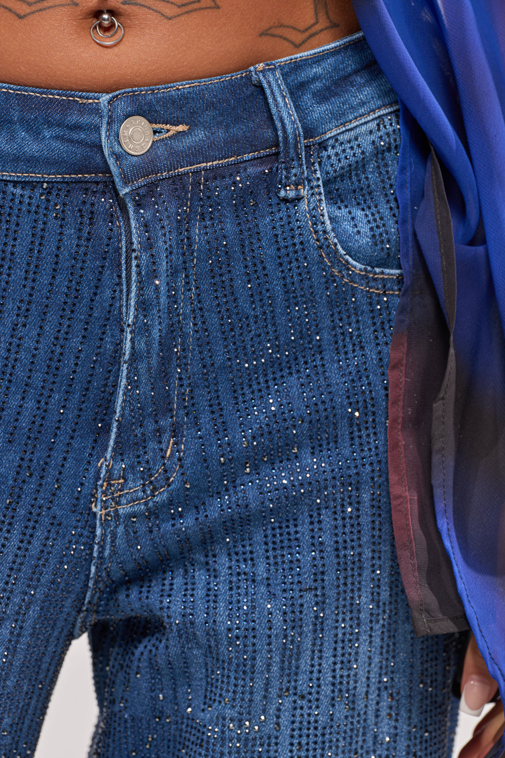 Close-up of a person wearing ITS ELECTRIC RHINESTONE WIDE LEG DENIM in blue, styled with a sheer blue and black top and a visible belly button piercing above the waistband. 6