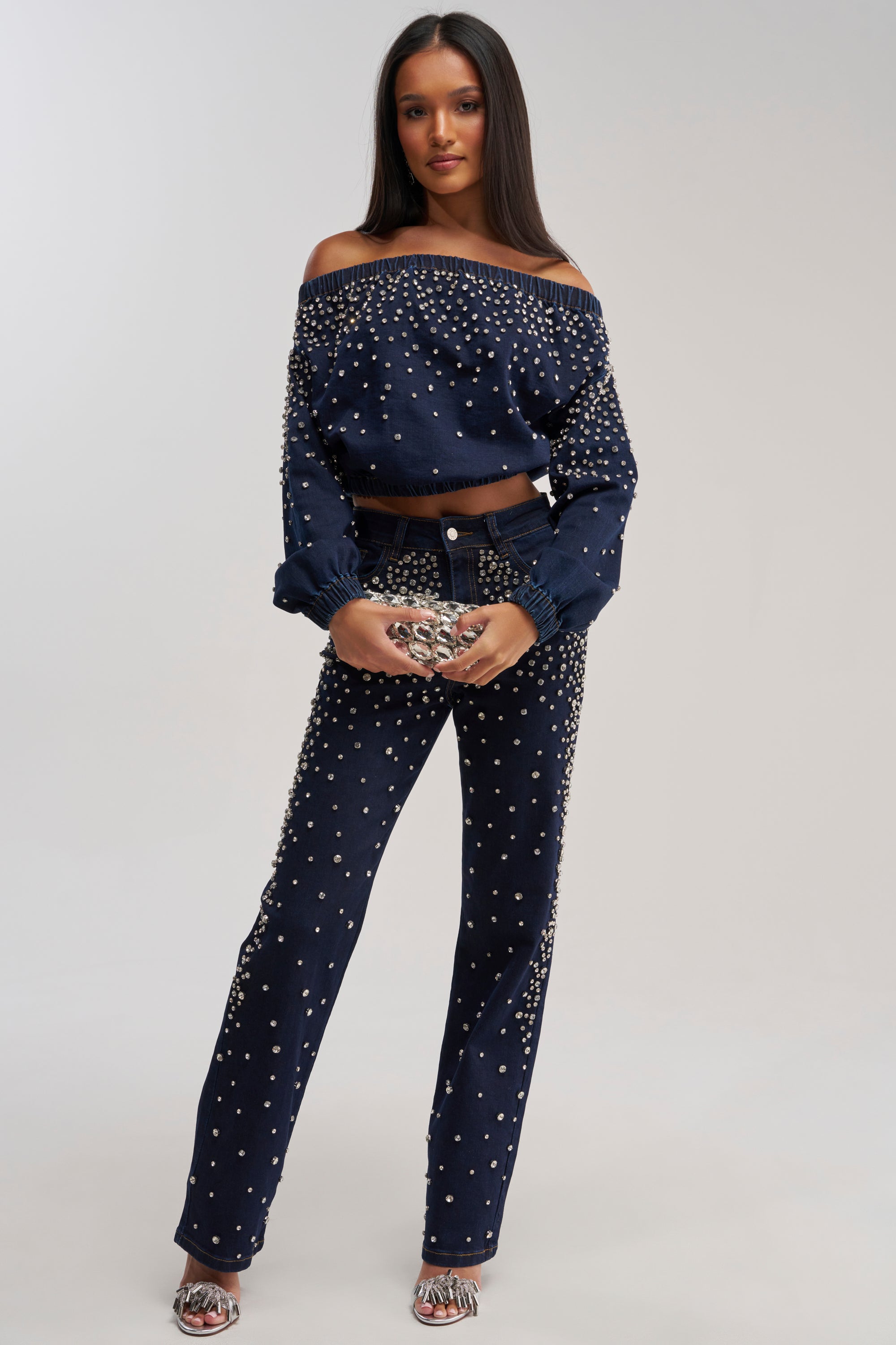 A woman faces forward in a SPARKLE SPARKLE EMBELLISHED DENIM SWEATSHIRT and matching studded pants, holding a small purse and wearing open-toe heeled sandals against a plain light background.