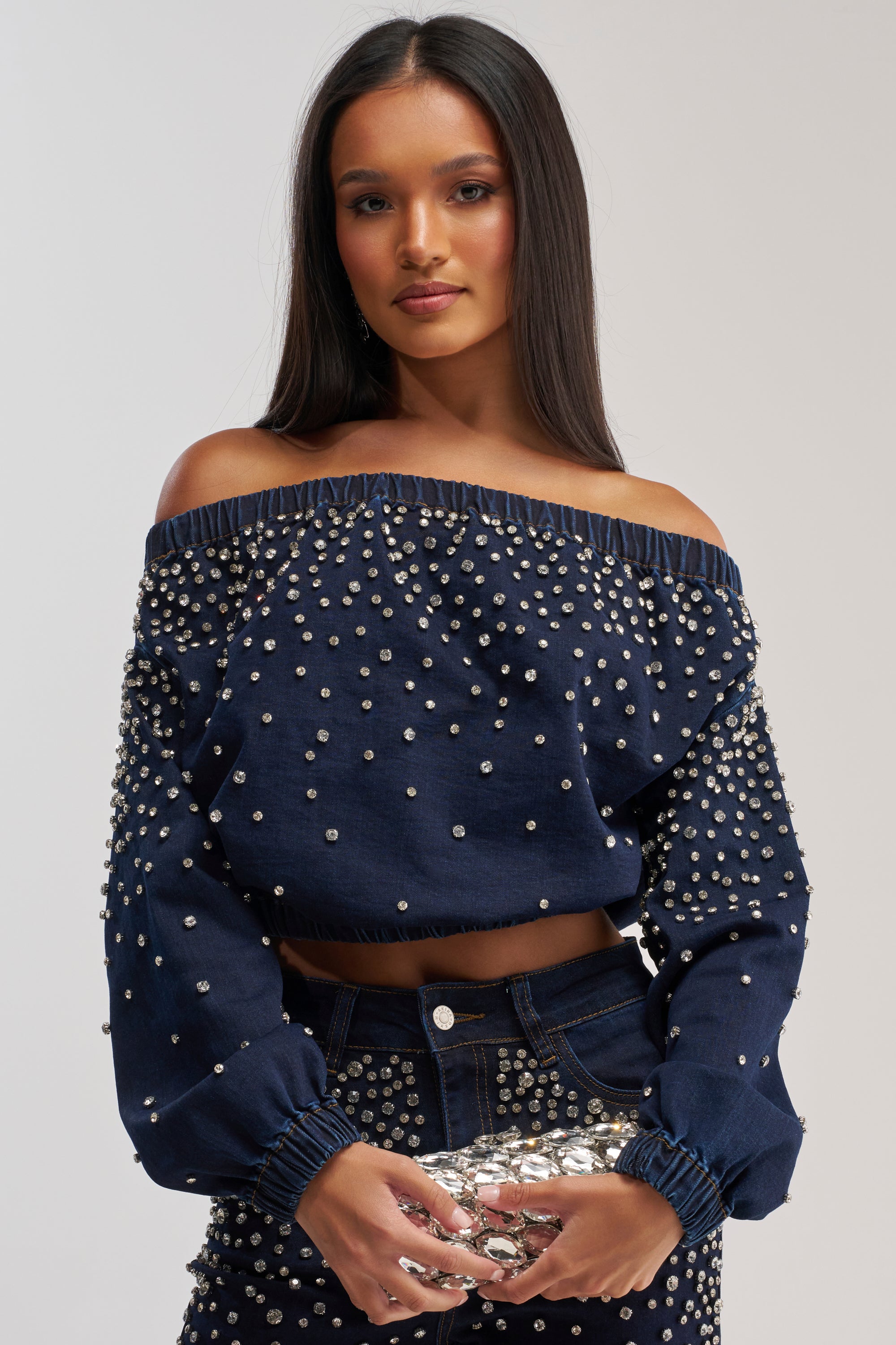Wearing the SPARKLE SPARKLE EMBELLISHED DENIM SWEATSHIRT and matching studded pants, a woman holds a shiny silver clutch while standing against a plain light background.