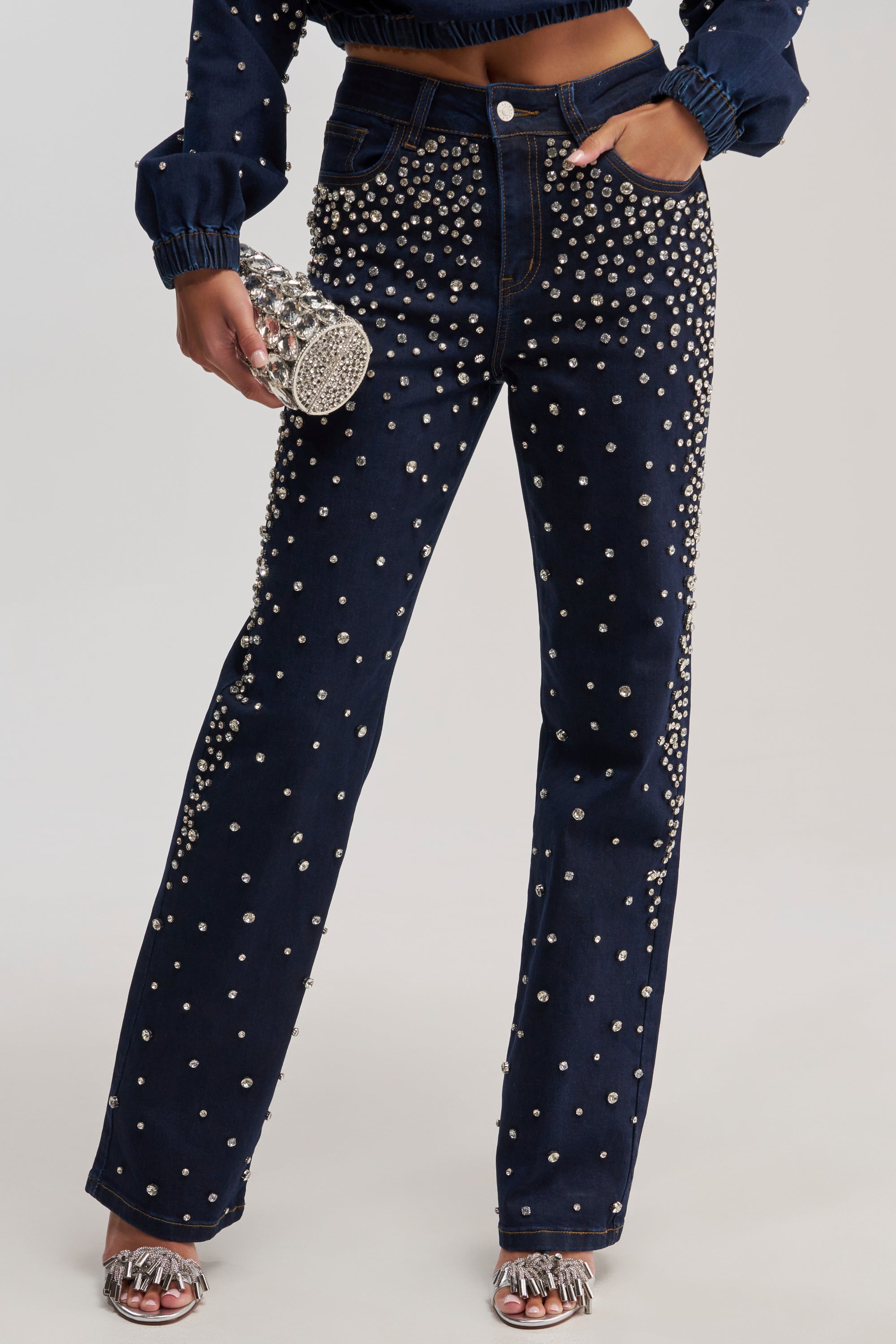 A person wears SPARKLE SPARKLE EMBELLISHED DENIM—high-waisted, dark wash jeans and a pearl-studded jacket—paired with silver sandals and a rhinestone clutch against a light gray background.