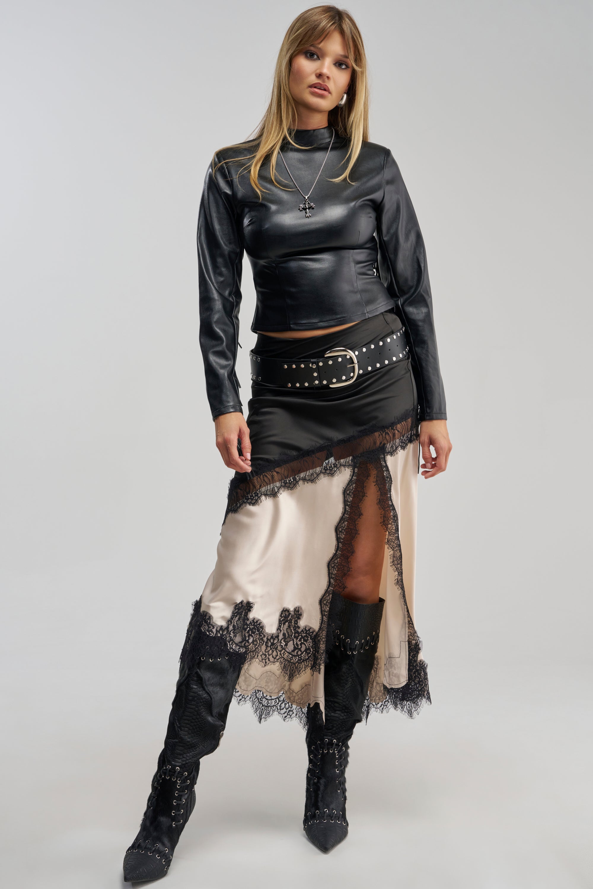 A woman stands facing forward against a plain background, styled in a black faux leather top, studded belt, and the SHINE BRIGHT SATIN MIDI SKIRT—featuring cream and black lace trim and a sheer lace-paneled side slit. Her look is completed with an asymmetrical black overlay and black lace-up boots.
