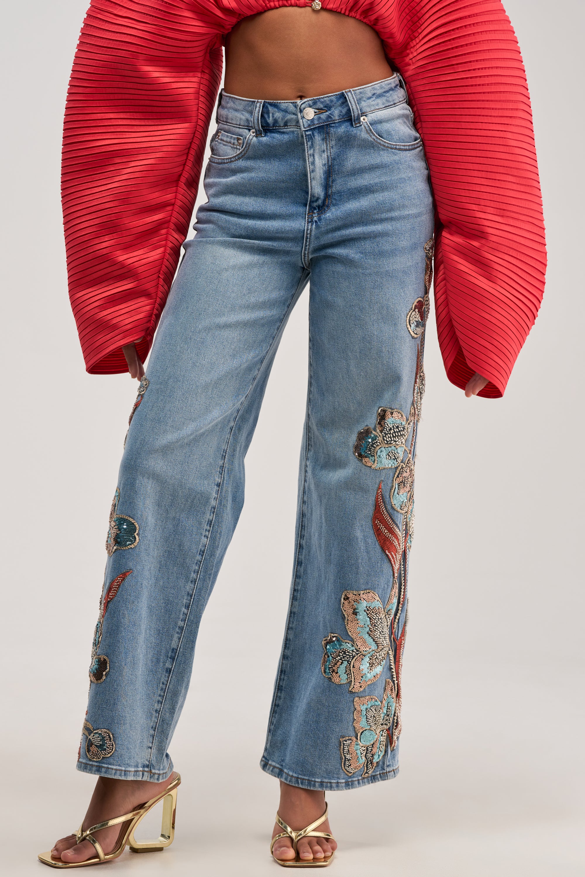 Wearing the PRETTY LIKE ME EMBELLISHED DENIM high-rise, wide-leg jeans with floral embroidery, paired with a red pleated long-sleeve top and gold heeled sandals.
