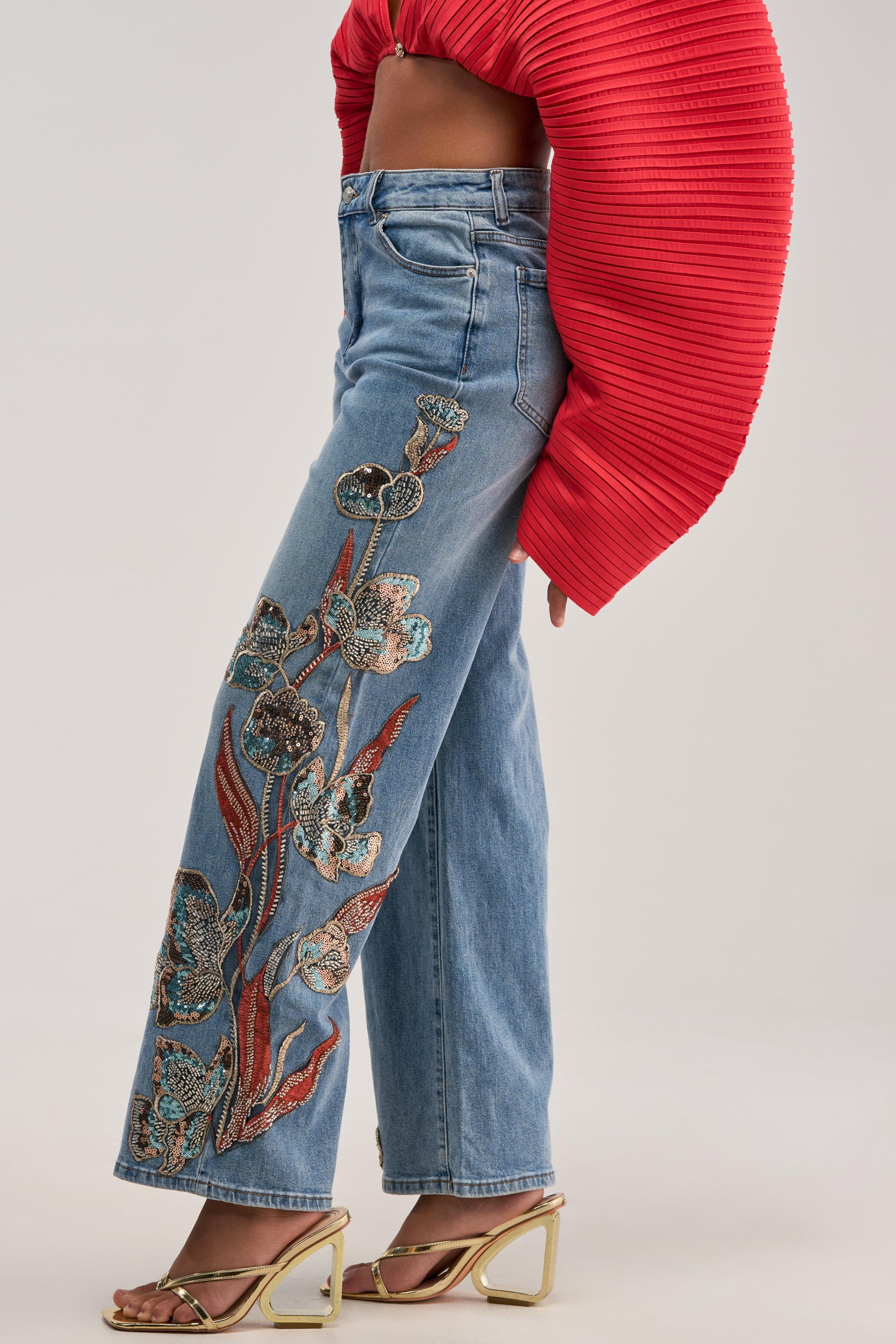 A person is wearing the PRETTY LIKE ME EMBELLISHED DENIM—high-rise jeans featuring floral embroidery with blue straight-leg denim on one leg—paired with a red textured long-sleeve crop top and gold open-toe heels. The background is plain and light-colored.
