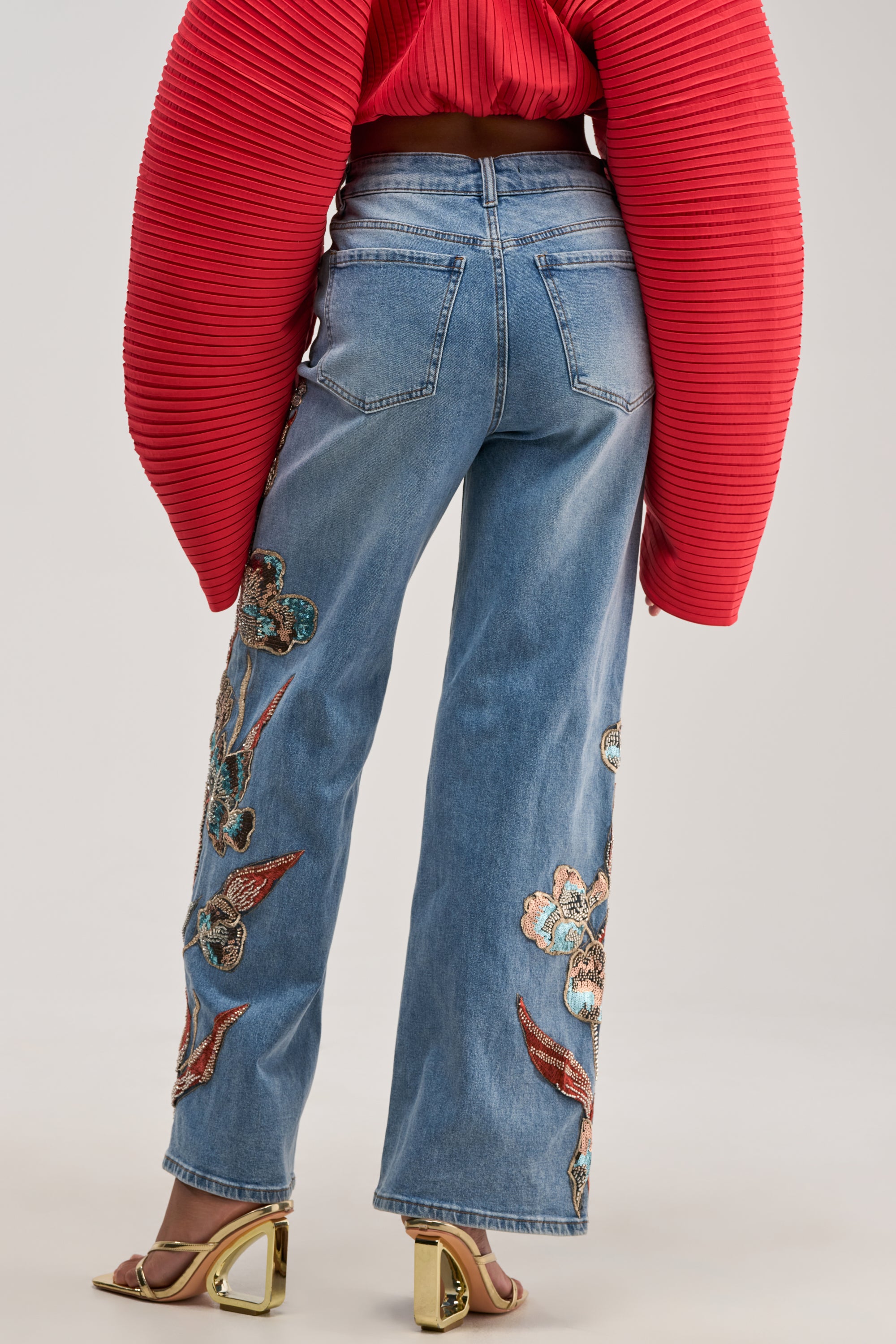 A person wearing a red textured top and the PRETTY LIKE ME EMBELLISHED DENIM stands facing away. They wear gold heels with clear block heels.
