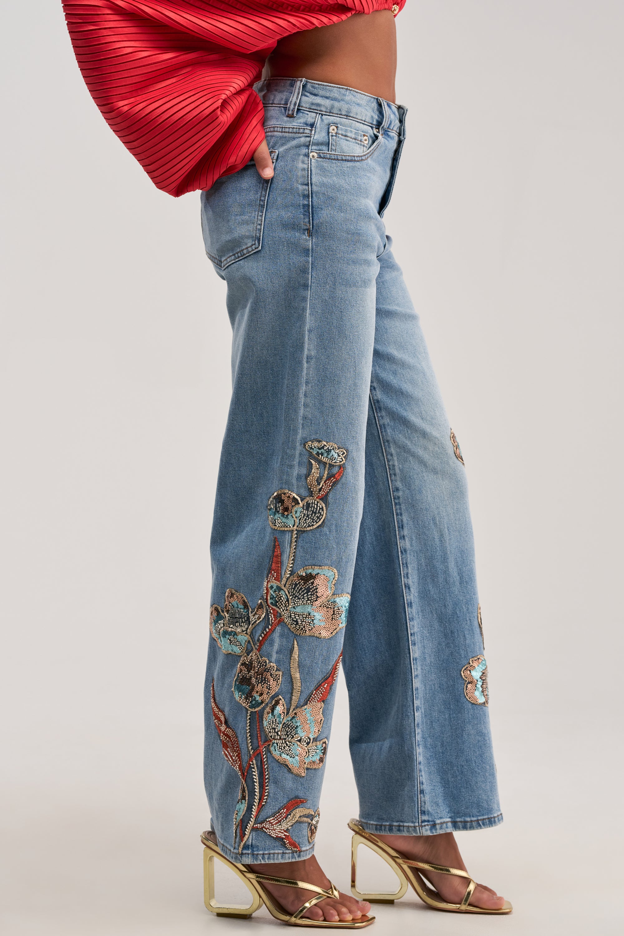 Wearing the PRETTY LIKE ME EMBELLISHED DENIM—light-wash straight leg jeans adorned with colorful floral embroidery—a person pairs them with a red textured top and gold high-heeled sandals, standing against a plain background.
