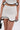 A person wears the CUTIE PIE RUFFLE HEM MINI SKIRT with a white textured crop top, both featuring wavy black trim and subtle ruffle hems. They hold a small black handbag against a plain white background.