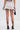 A person stands with their back to the camera, wearing the CUTIE PIE RUFFLE HEM MINI SKIRT in white with black trim, paired with black strappy high-heeled sandals and holding a small black handbag.