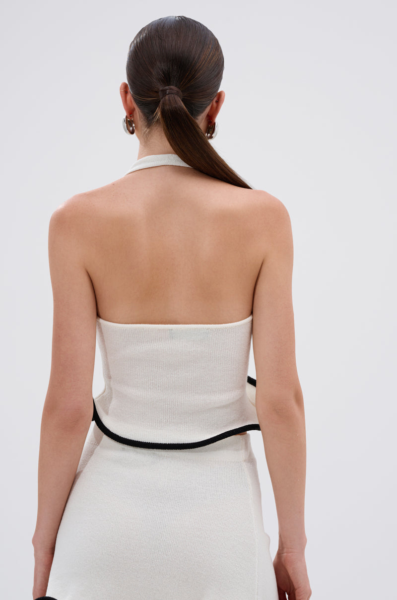 A woman with straight brown hair in a low ponytail is seen from behind wearing the CUTIE PIE RUFFLE HEM BLOUSE, a strapless white top with black trim made from stretchy knit material. She pairs it with a matching white skirt; background is plain and light. 3