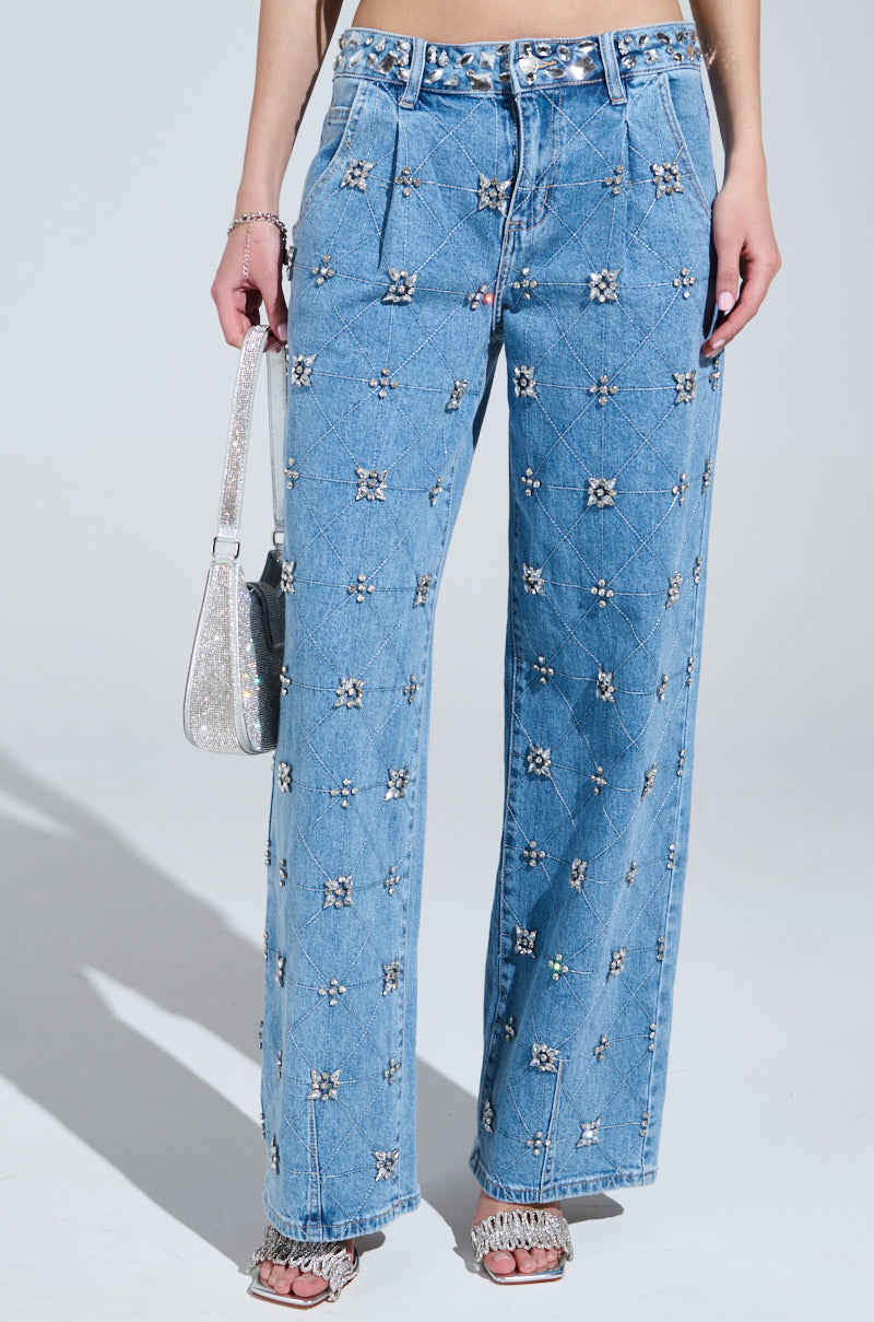 Wearing TAKE ME OUT RHINESTONE DENIM JEANS adorned with sparkling silver rhinestones, a person holds a glittery silver handbag and sports open-toed fringe heels, standing out against a simple light gray background.