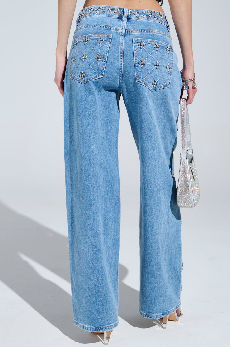 A person faces away wearing TAKE ME OUT RHINESTONE DENIM JEANS, featuring jeweled flower designs on the back pockets. They're holding a sparkling silver handbag and wear white heeled shoes.