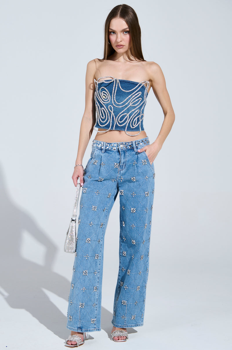 A woman wears the TAKE ME OUT RHINESTONE DENIM JEANS—high-rise straight-leg denim accented with sparkling silver rhinestones—paired with a strapless swirl-patterned top, open-toe heels, and a small silver bag.