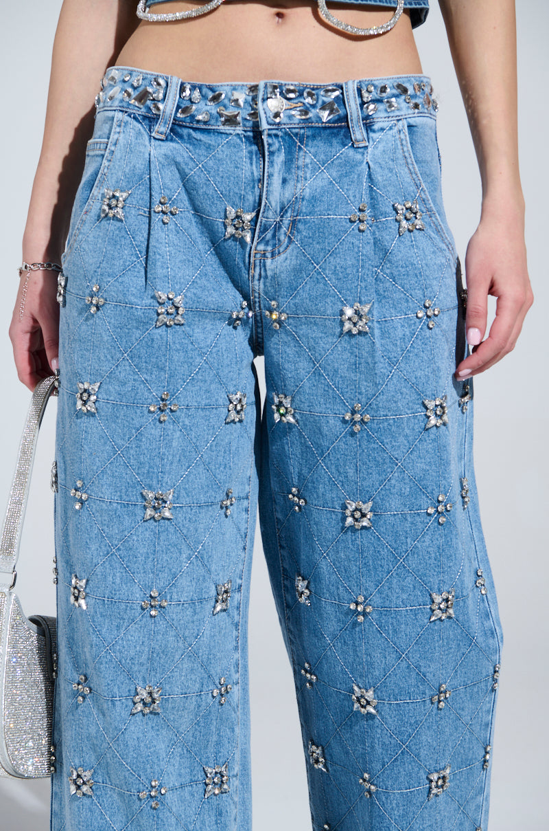 Close-up of a person wearing TAKE ME OUT RHINESTONE DENIM JEANS, featuring sparkling silver rhinestones in a diamond pattern. These high-rise straight-leg jeans are styled with a glittery silver belt and matching handbag.
