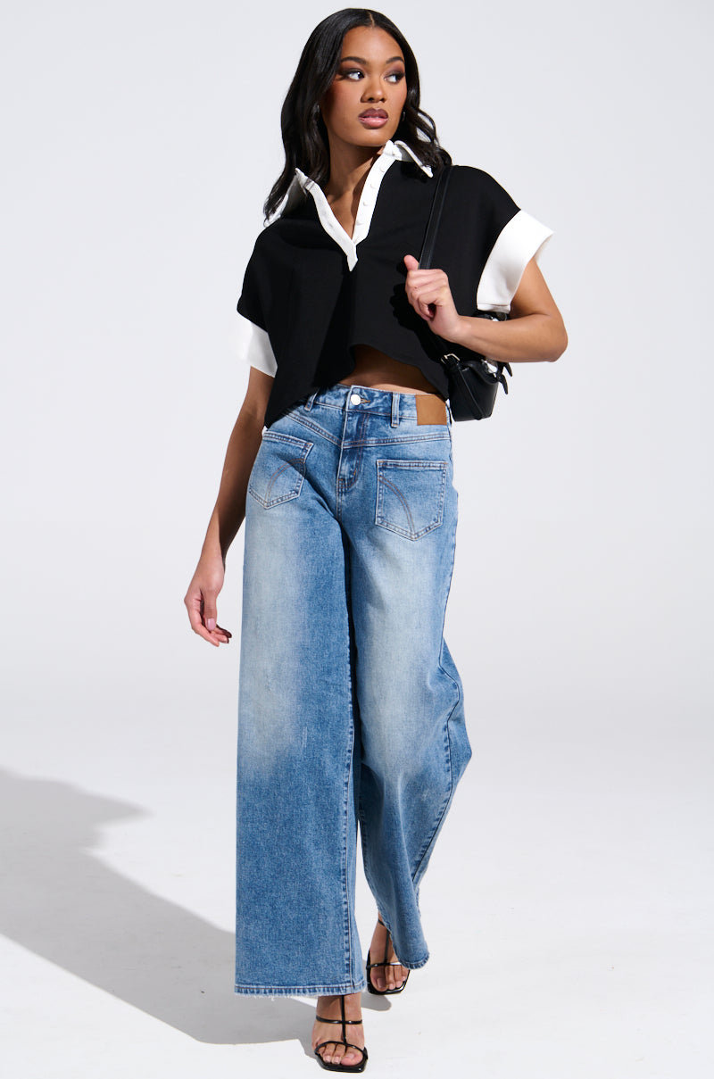 A woman poses confidently against a light gray backdrop in a black cropped top over a white shirt, flaunting her high-rise REVERSE REVERSE MEDIUM WASH JEANS. She accessorizes with black sandals and a small black shoulder bag while showcasing the jeans' wide-leg silhouette.